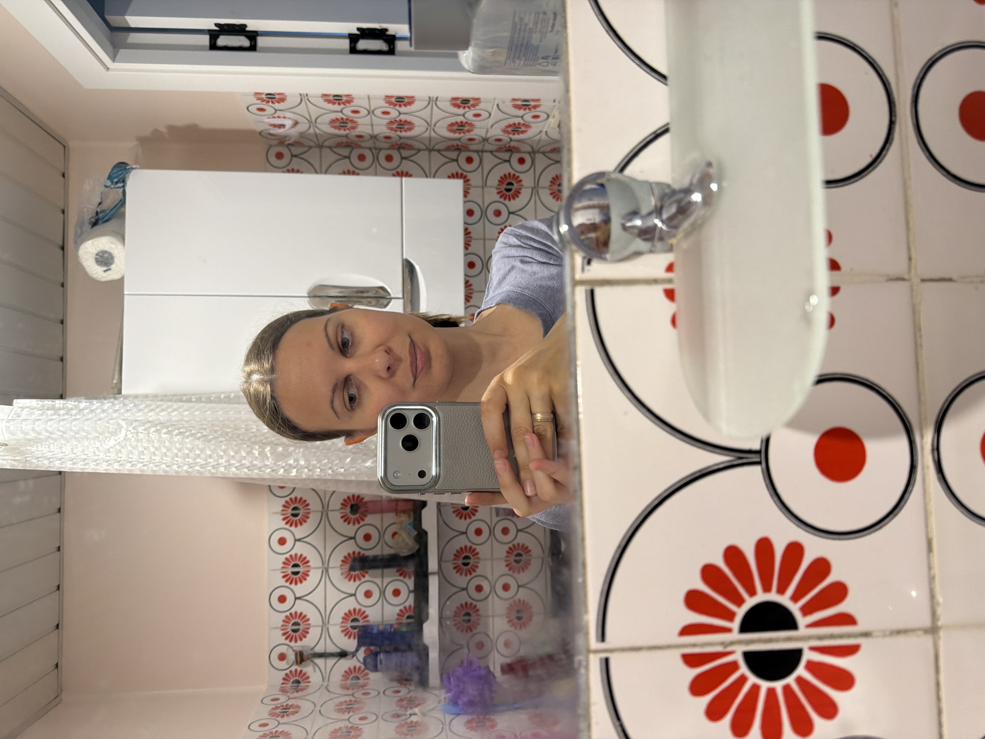 The image shows a woman taking a selfie in a bathroom. She is holding a gray iPhone in her right hand and looking at the camera. She is wearing a gray t-shirt and has a ring on her left ring finger. The bathroom has pink walls and white tiled walls with a red floral pattern. There is a white cabinet with a roll of toilet paper on top, a shower curtain, and a sink with a chrome faucet. The image appears to have been taken in a residential bathroom. The provided metadata does not contain information about the specific location within Izhevsk, Russia.