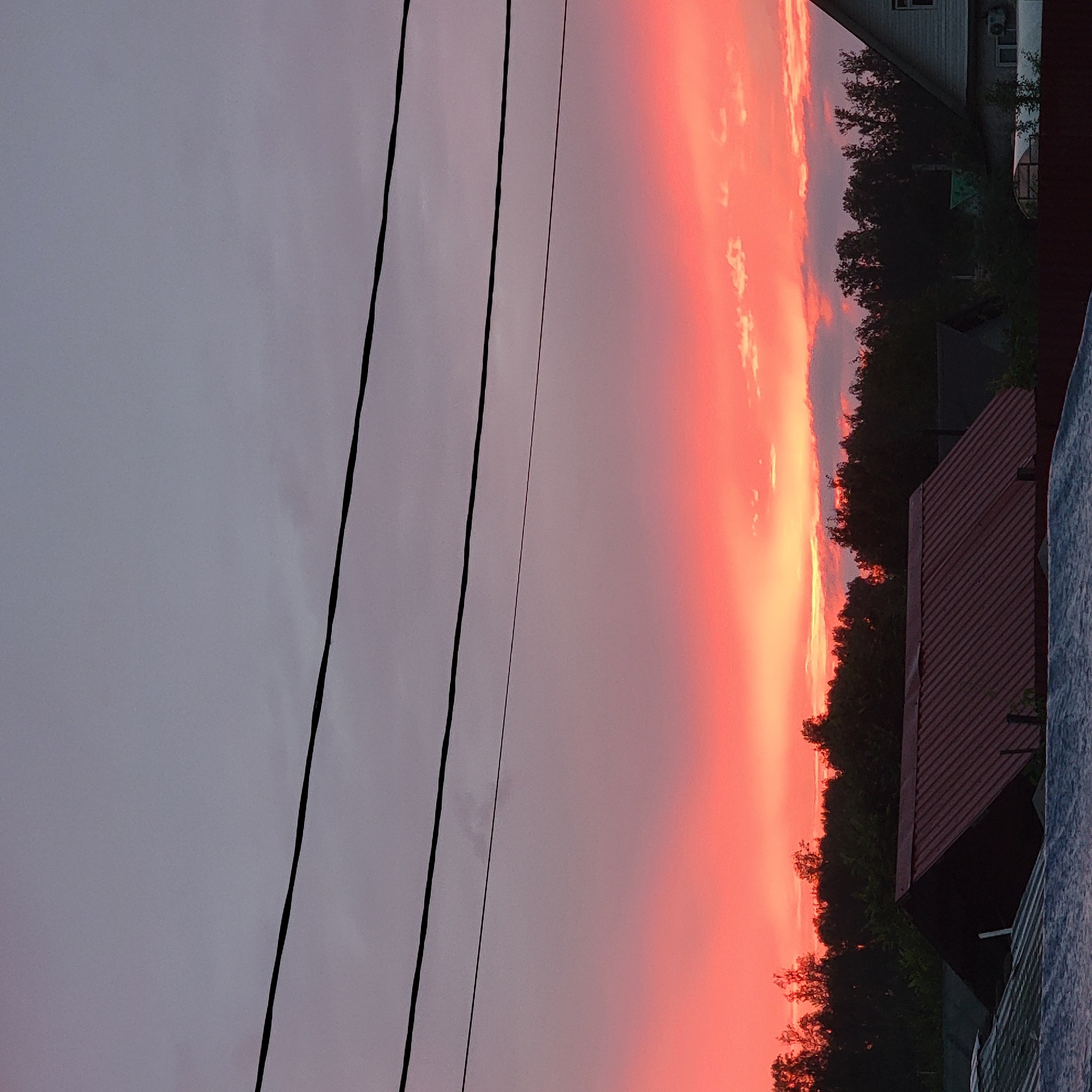 This is a photograph of a sunset over a rural landscape, likely on the outskirts of Iglino, Russia. The sky is dominated by vibrant hues of pink and orange, transitioning to a muted grey at the top. Three dark power lines traverse the sky horizontally, from left to right. Below the sunset, a dense line of dark green trees forms a silhouette against the colorful sky. In the foreground, several rooftops are visible, including a prominent dark red metal roof on a building with a gabled roof. To the right, a portion of a light-colored building with a window and an air conditioning unit is visible. The overall scene depicts a peaceful evening in a rural or semi-rural setting.