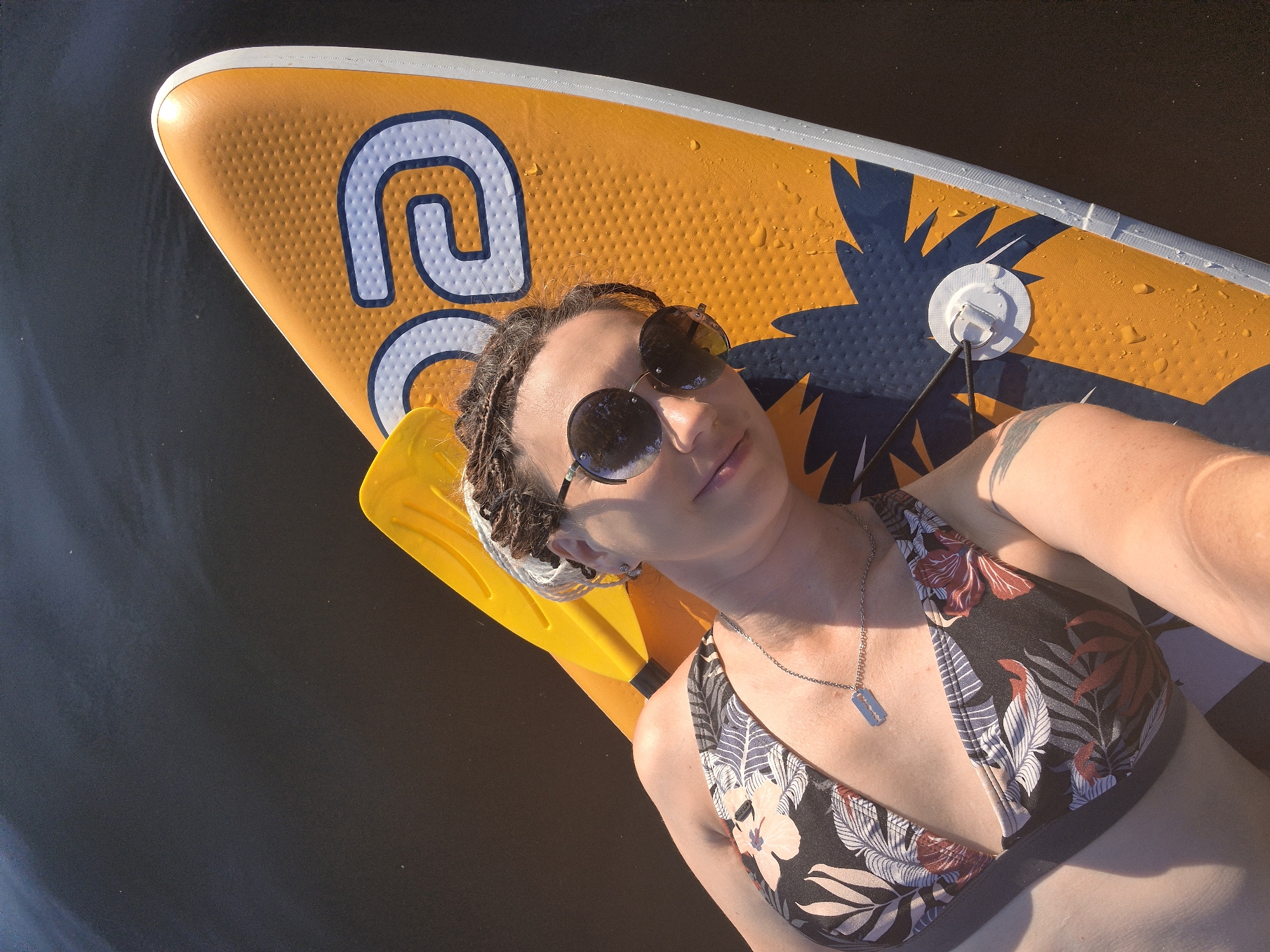 A woman with dark, braided hair and round sunglasses lies on her back on an orange stand-up paddleboard in dark water. She is wearing a floral bikini top and a silver necklace with a razor blade pendant. A yellow paddle is positioned behind her head. The paddleboard has a blue and white logo and a palm tree design. The scene appears to be outdoors on a sunny day.