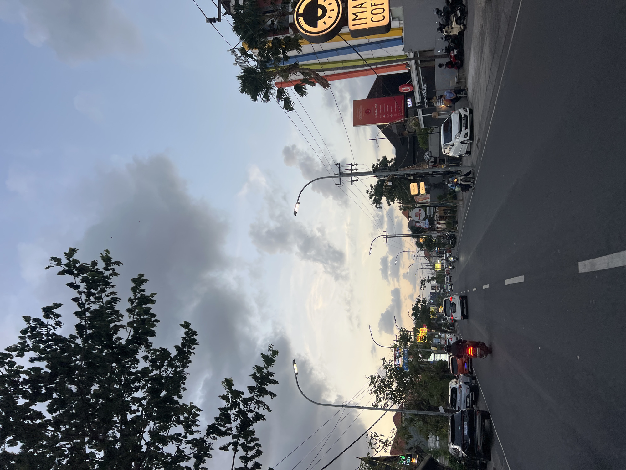 The image depicts a street scene in Kuta, Indonesia, during twilight. The sky is a mix of gray clouds and pale blue, with hints of sunset colors near the horizon. A large tree with dark leaves overhangs the left side of the frame. The road is asphalt, with white dashed lines marking the lanes.
Several vehicles are present: a black SUV parked on the left, a white SUV parked further down, and a white hatchback in the right lane. A motorcycle with a rider is moving away from the camera in the right lane. Several other motorcycles are parked on the right side of the road.
On the right side of the street, a building with colorful vertical stripes and a large circular sign with a logo is visible. Text on the sign reads "IMAN COF". Other businesses with illuminated signs are present further down the street. Several people are visible on the sidewalk, some sitting, others standing or walking. Streetlights are illuminated along the road. The overall atmosphere suggests a busy street at the end of the day.