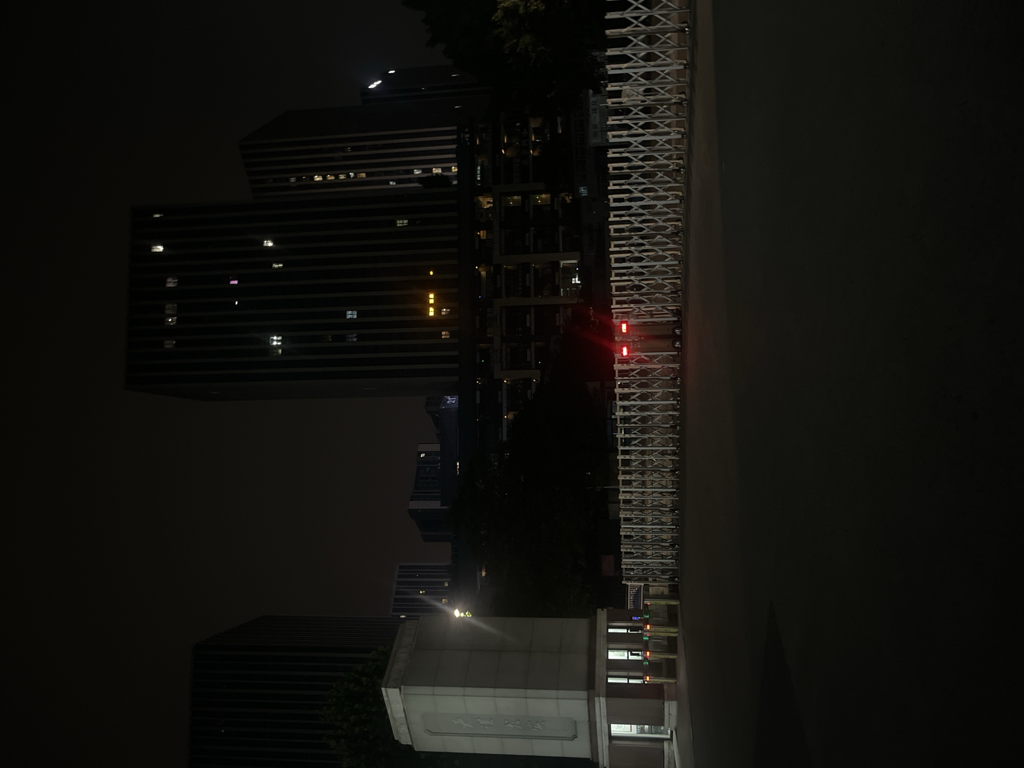 The image depicts a nighttime scene outside a gated entrance in Yuzhong District, China. In the foreground, a dark asphalt surface leads to a series of collapsible metal gates. Red traffic lights are visible, indicating a stop or warning. To the left of the gates is a white stone structure that appears to be a guardhouse or entrance monument, with illuminated windows and text in Chinese characters on its surface. Behind the gates and guardhouse, several modern high-rise buildings rise into the dark sky. These buildings have vertical lines and illuminated windows, suggesting office spaces or residences. Trees and foliage are present between the entrance and the buildings, partially obscuring the lower levels of the structures. The overall atmosphere is dark and urban, with artificial lighting from the buildings and entrance creating a contrast with the night sky.