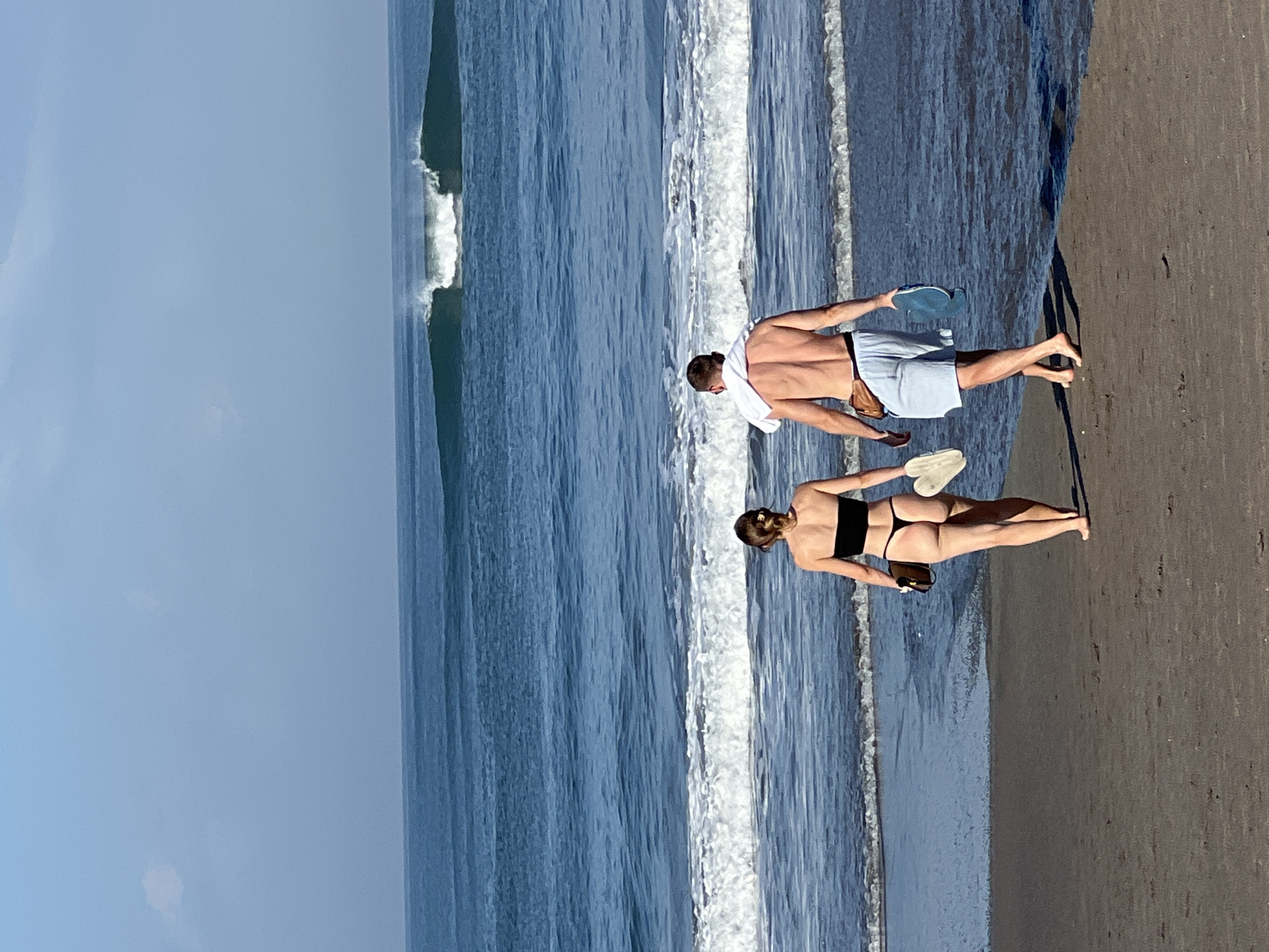 A man and woman walk away from the camera on a dark sand beach towards the ocean in North Kuta, Indonesia. The woman wears a black strapless bikini and carries a brown bag and a white flip-flop. The man wears light blue swim trunks, a brown fanny pack, and carries a blue flip-flop. A white towel is draped over his shoulders. The ocean has moderate waves, with a large breaking wave in the distance. The sky is clear and blue with a few faint clouds. The sun appears to be high in the sky, indicating it is likely midday.