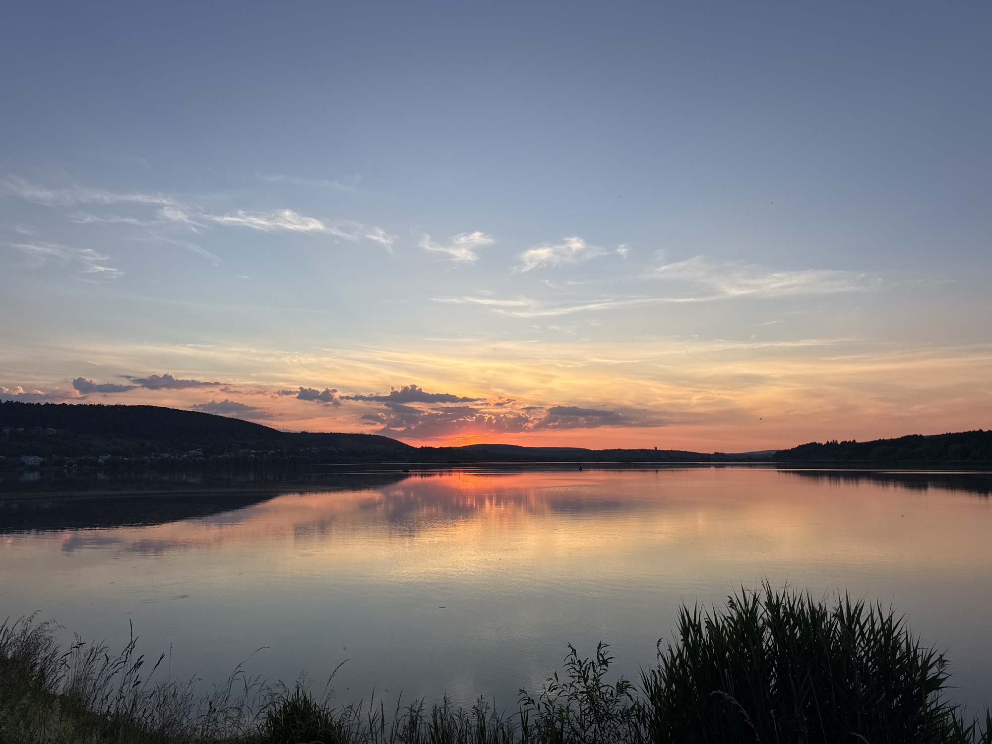 The image depicts a sunset over a wide body of water, likely a river or lake, in Berezhany, Ukraine. The sky is a gradient of blue at the top, transitioning to hues of orange, pink, and yellow near the horizon where the sun is setting behind rolling hills. Wispy clouds are scattered across the sky, catching the warm light of the sunset. The water reflects the colors of the sky and the silhouetted landscape, creating a mirror image. On the left side of the image, hills covered in trees and some scattered buildings are visible, with their dark silhouettes contrasting against the bright sky. The foreground is occupied by tall grasses and reeds, slightly out of focus, which frame the scene and suggest a natural embankment. There are no people or specific actions visible in the image, rendering a tranquil and serene natural landscape. The scene is devoid of any specific urban elements, emphasizing the natural beauty of the sunset.