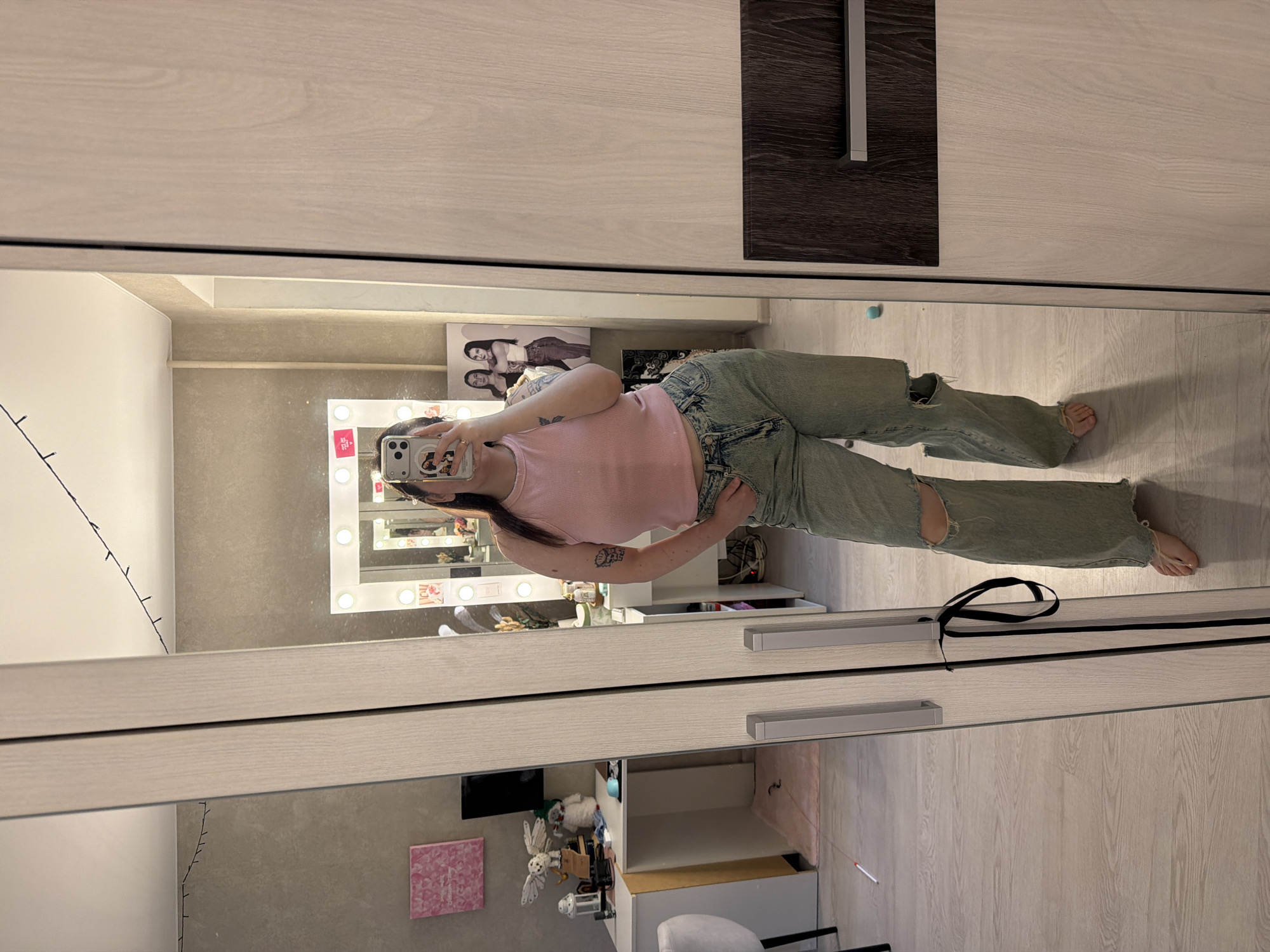 A woman in a pink tank top and ripped jeans is taking a selfie in front of a wardrobe mirror in a room in Chaikovsky, Russia. The room contains a vanity with a lighted mirror, various cosmetics, and decorative items on shelves. The wardrobe has sliding doors, one light wood and one dark wood. There is a poster on the wall above the vanity. A black ribbon lies on the floor near the mirror.