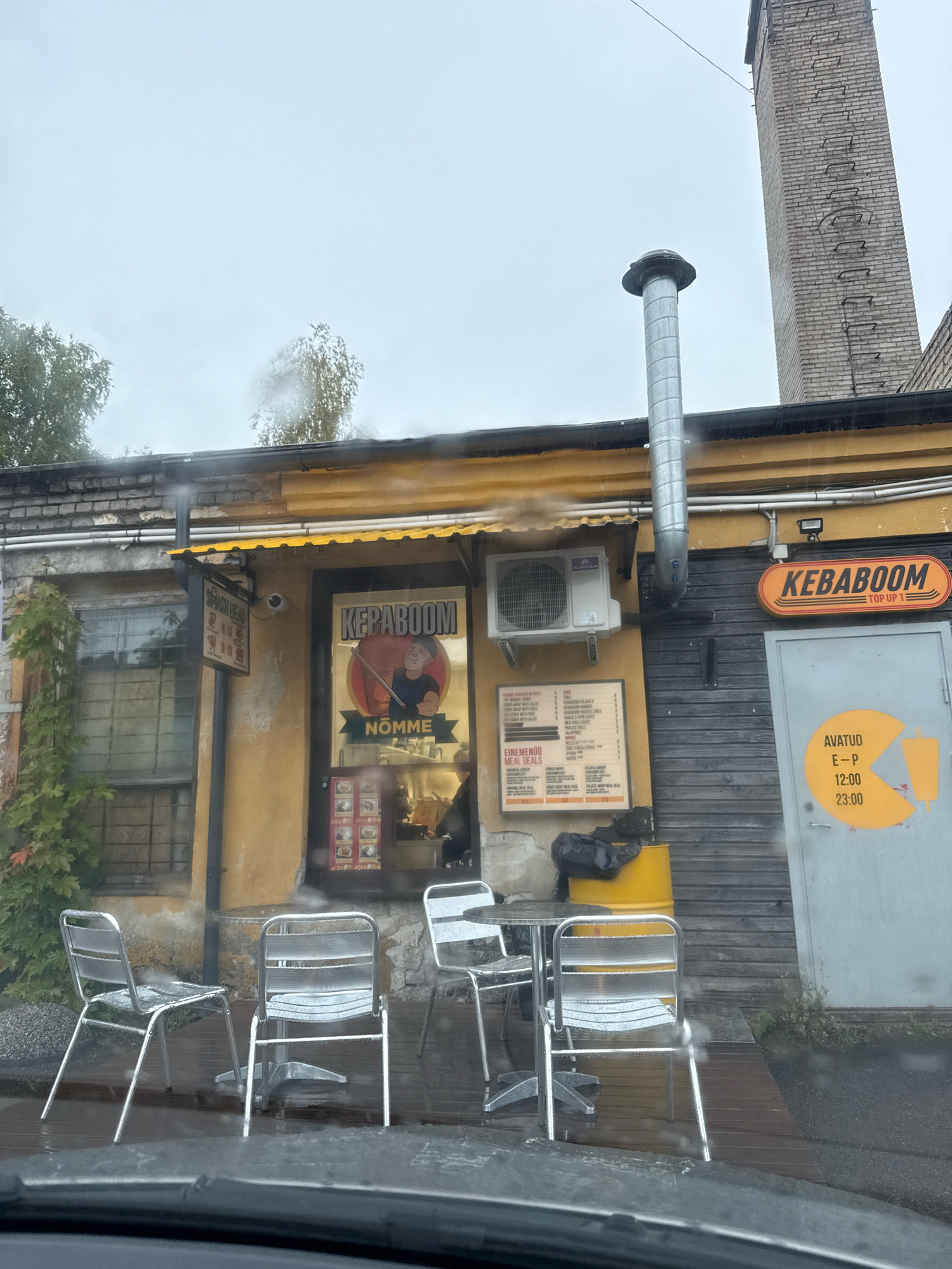 This is an outdoor, eye-level shot of a small food establishment in Tallinn, Estonia, under a cloudy sky with visible raindrops on the camera lens. The building's facade is a weathered yellow with exposed brick at the top and a gray wooden plank section. A yellow awning extends over the main entrance.
A sign above the door reads "KEBABOOM" in orange lettering on an orange oval background. To the left of the entrance, a window displays a menu with small images of food items. Another sign, a framed menu, is mounted on the wall to the left of the window, listing various food items and prices. An outdoor air conditioning unit is mounted on the wall above this menu.
In front of the establishment, there are three empty, metallic chairs and one table, all appearing wet from the rain. A large, yellow barrel sits to the right of the entrance, behind some of the chairs. A metal pipe extends vertically from the roof near the air conditioning unit, and a large brick chimney is visible to the right of the building.
A gray door on the far right has an orange circular sign with what appears to be opening hours: "AVATUD E-P 12:00 23:00" (Open Monday-Sunday 12:00 PM to 11:00 PM). The ground in front of the establishment is a wooden deck, also appearing wet. There is dense greenery on the left side of the building. The overall impression is of a humble, possibly old, and operational food vendor.