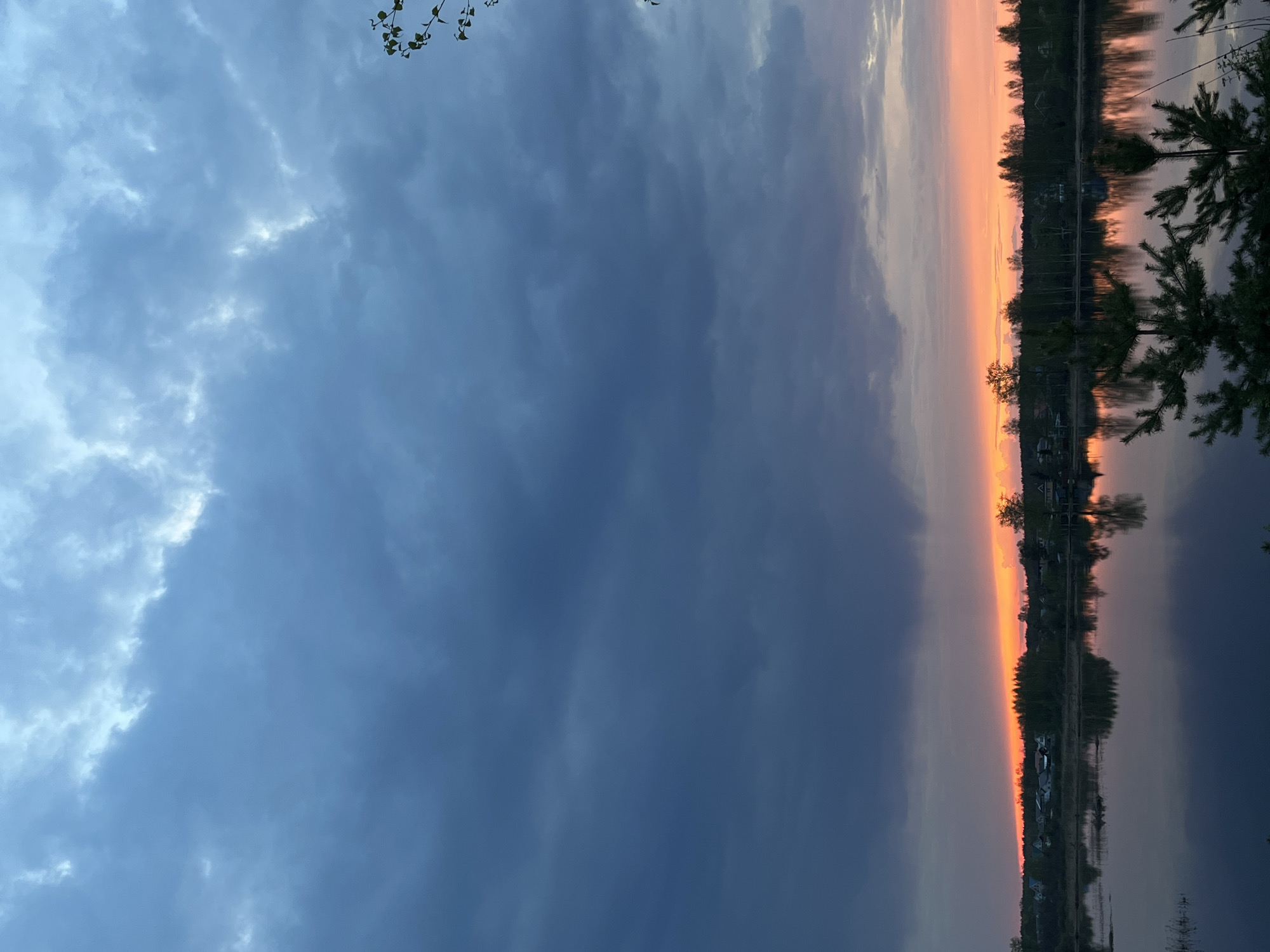 The image shows a sunset scene in Bol'shoye Kozino, Russia. The sky is divided between dark, stormy clouds on the left and a vibrant orange and pink sunset on the right. The sunset reflects on the surface of a body of water, likely a lake or river. Trees line the horizon on the opposite shore, silhouetted against the sunset colors. Some vegetation is visible in the foreground as well. There are some objects that look like buildings in the background.
