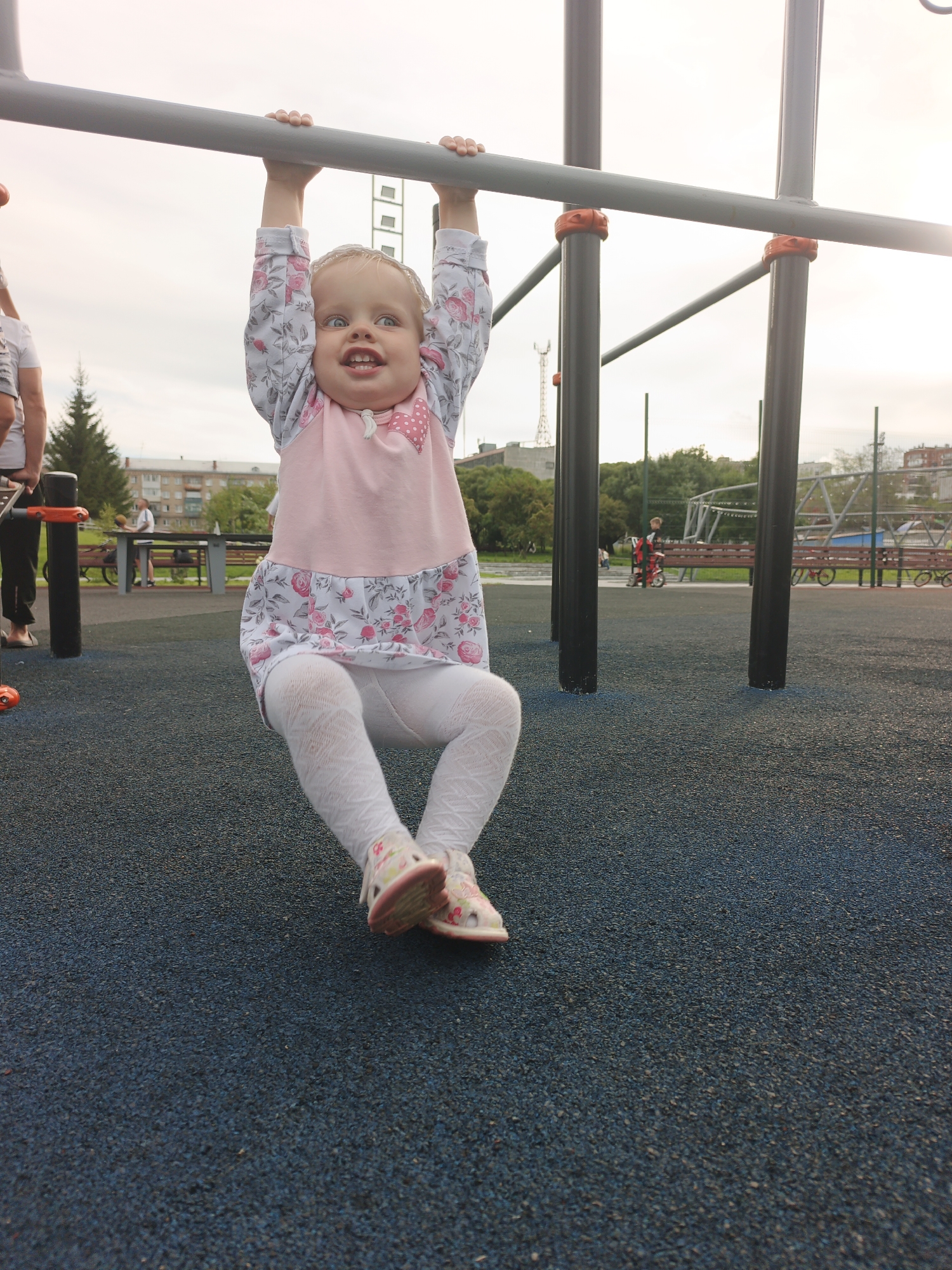A young Caucasian girl with blonde hair and blue eyes is hanging from a horizontal bar on an outdoor playground. She is wearing a pink and white floral dress, white textured tights, and pink and white sandals. Her mouth is open, and she appears to be smiling or laughing. The playground has a dark blue rubberized surface. In the background, there are other playground structures, trees, buildings, and people. The scene is set in the city of Zlatoust, Russia, indicated by the presence of specific architectural styles and vegetation common to the region. The lighting suggests it is daytime, likely late afternoon due to the soft light.