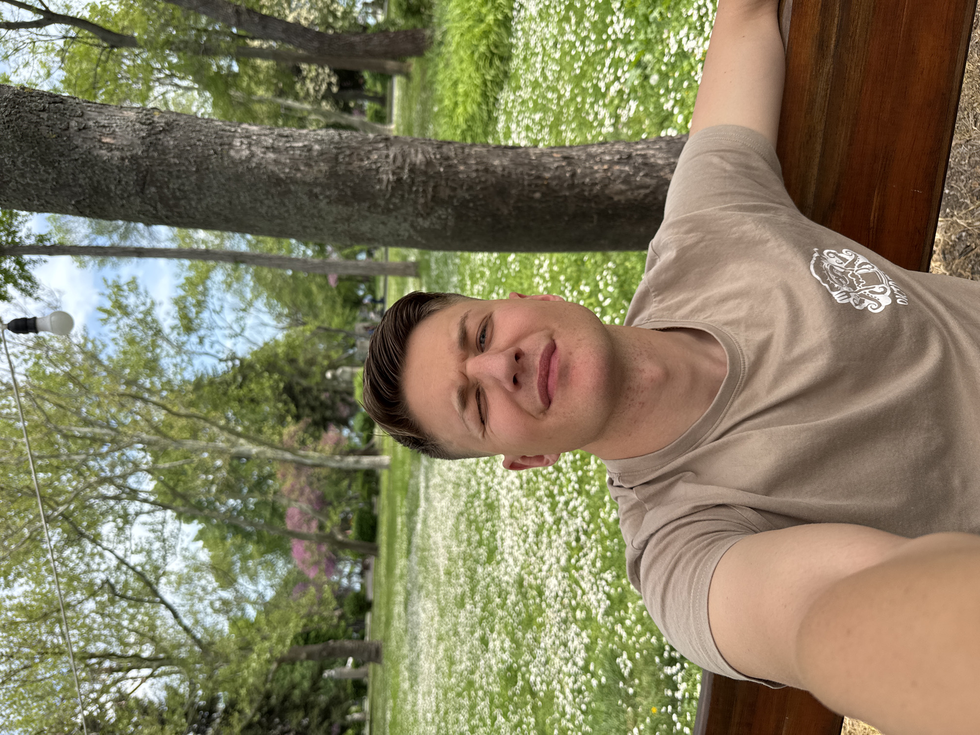 A man is sitting on a wooden park bench in a grassy area filled with white flowers. The man is wearing a brown t-shirt with a white design on the chest. He is looking at the camera and winking. There is a large tree trunk behind him and other trees and sky visible in the background. The scene is possibly set in a park in Burgas, Bulgaria.