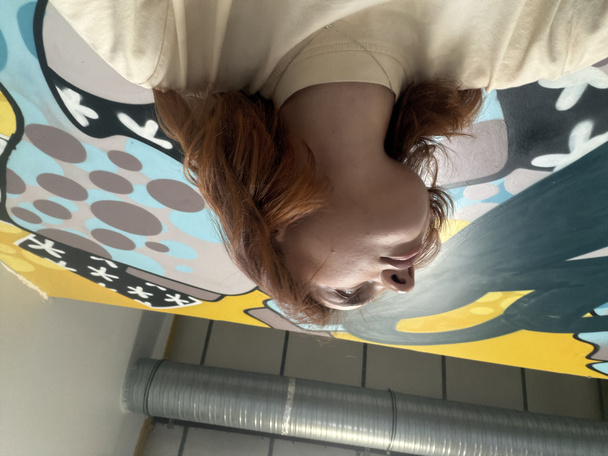 The image shows a person with reddish-brown hair, wearing a light-colored t-shirt, leaning against a large, colorful mural. The mural features abstract shapes in yellow, blue, gray, and black, with some white accents. To the left of the person and mural, a large, silver, corrugated pipe is visible, attached to a wall. The background appears to be an indoor space, possibly a studio or workshop, with muted grey walls. The lighting suggests it is daytime. There are no discernible elements in the image that would indicate the location is Strasbourg, France.