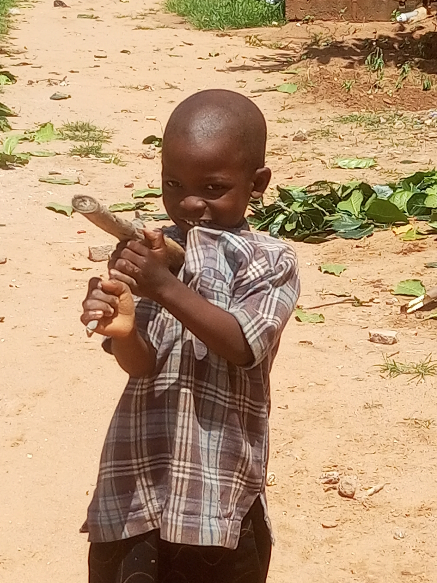 A young boy stands outdoors in a sandy area. He is holding a long, narrow object, possibly a stick or a piece of wood, horizontally in front of him, gripping it with both hands. The boy is wearing a plaid shirt. The ground is covered in sand and dry leaves, with some patches of green vegetation visible in the background. The overall setting suggests an unpaved area, potentially on the outskirts of a settlement. This scene is located in the city of Tempure, Nigeria.