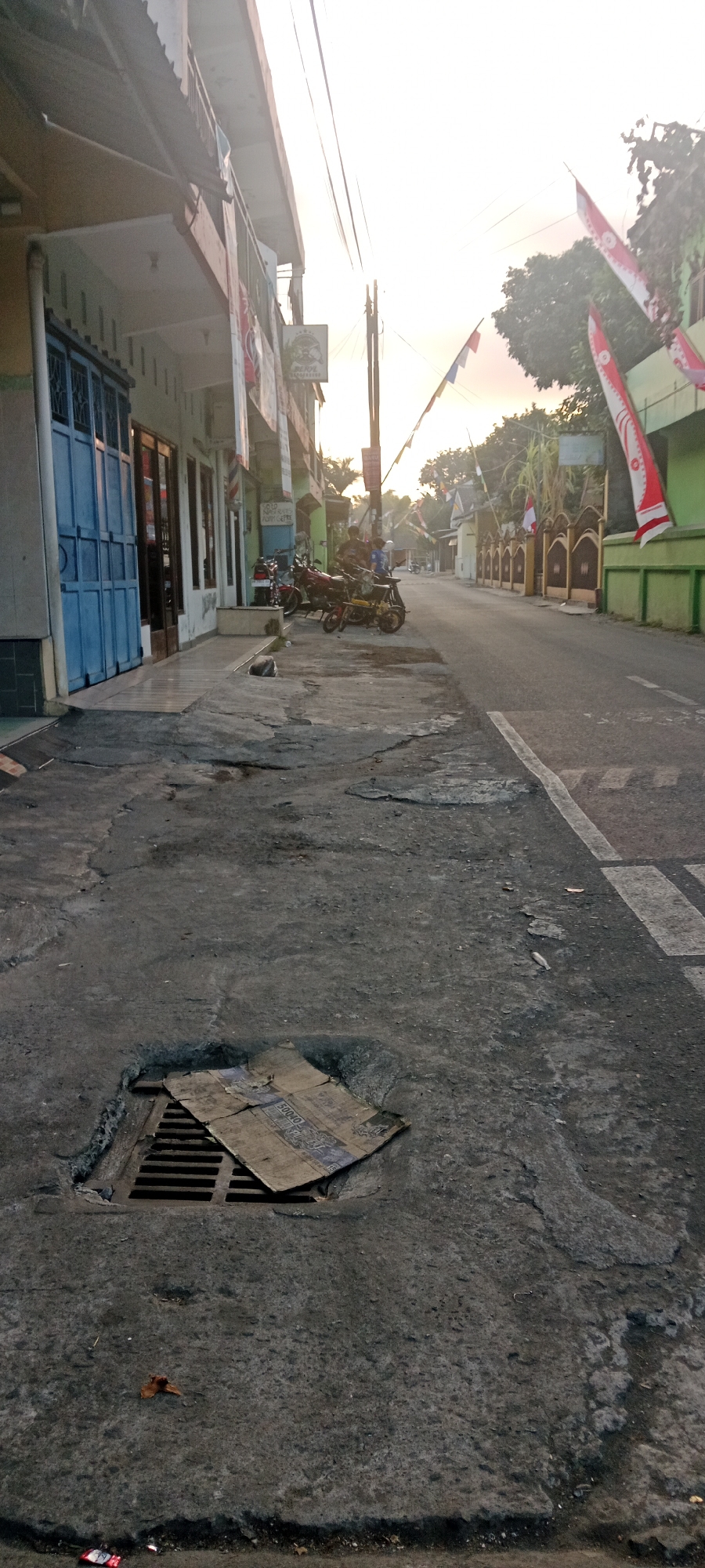 In the early morning light in Maguwoharjo, Indonesia, a narrow street is lined with shops and houses. The pavement is cracked and uneven, with a drain grate covered by a piece of cardboard in the foreground. Several motorcycles are parked along the sidewalk in front of the buildings. Indonesian flags and banners hang from poles along the street, suggesting a local celebration or event. The sun is low on the horizon, casting long shadows and creating a warm, hazy atmosphere.