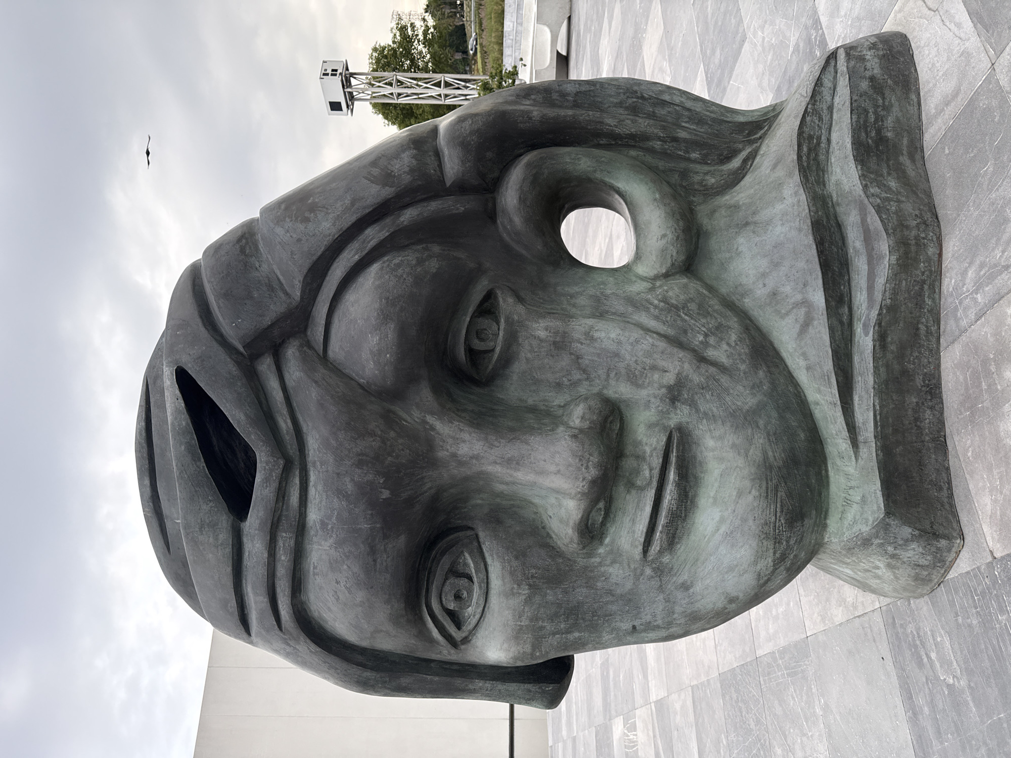 The image shows a bronze sculpture of a human face, with stylized features and openings where the eyes and forehead would be. The sculpture is situated on a light grey tiled surface. In the background, there is a white building, a metal structure possibly a lifeguard tower, trees, and an overcast sky. A bird is visible flying in the upper left of the image. The sculpture is located in the city of San Bernardino Tlaxcalancingo, Mexico.