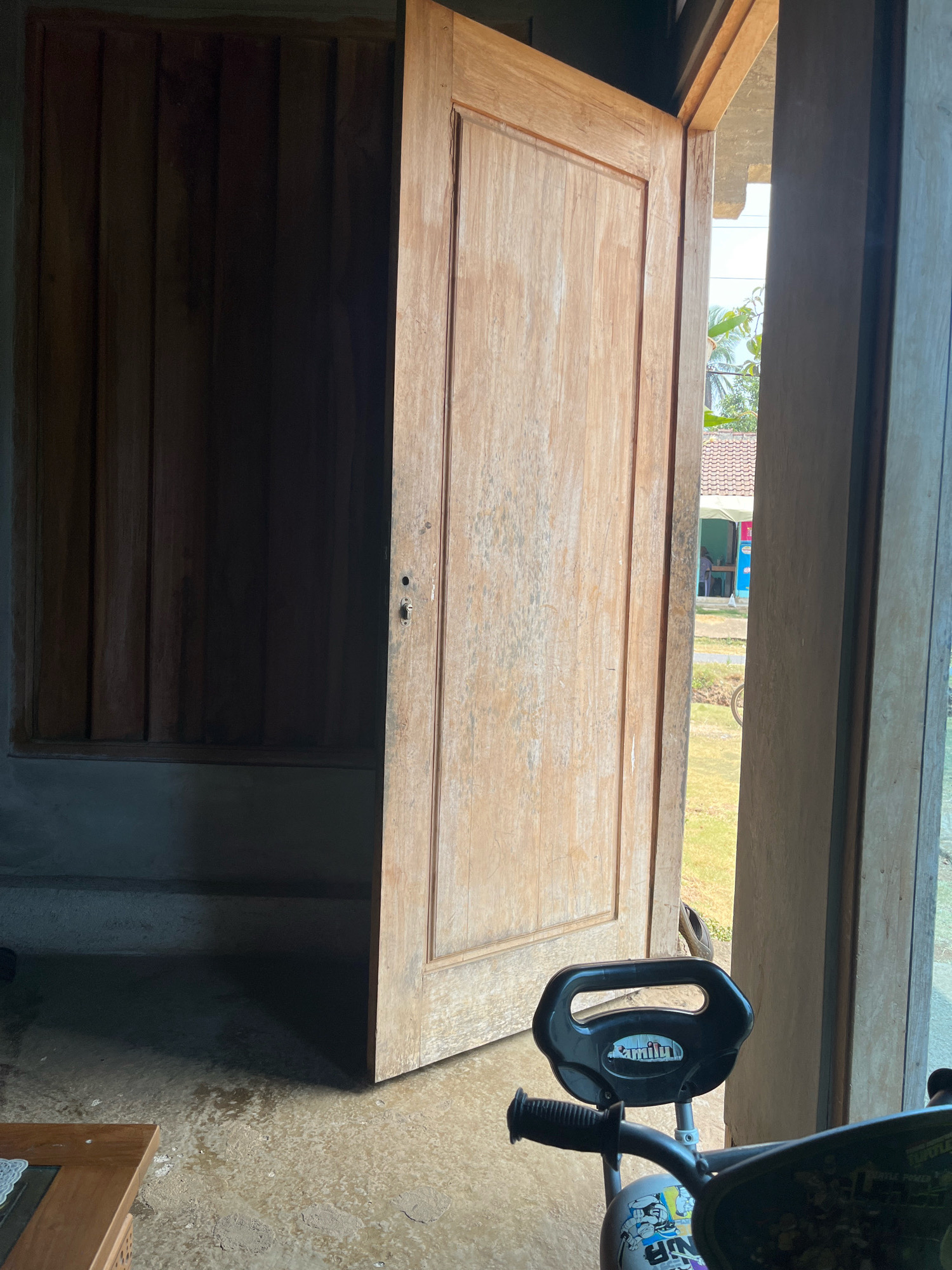 An open wooden door reveals a view of an outdoor scene in Sinar Mulyo, Indonesia. The door, made of light-colored wood with a visible grain, is partially open, allowing a sliver of sunlight to illuminate the interior space. To the left of the door is a dark wooden paneling. In the foreground, partially obscuring the view through the doorway, is a black plastic handlebar of a child's tricycle, with the word "Family" visible on a sticker. Beyond the open door, a green grassy area is visible, with a tiled roof of a building in the background and some trees and foliage. The ground inside the doorway appears to be a concrete floor, with some patches of dirt and moisture.
