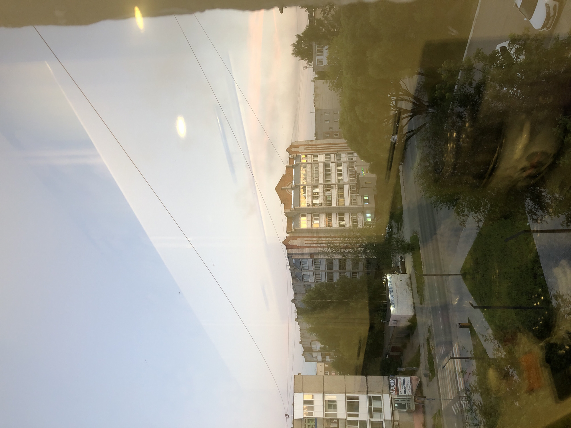 The image presents a view from a window in the city of Solikamsk, Russia, looking out at an urban landscape during twilight. In the foreground, reflections on the glass obscure parts of the scene, creating a yellowish tint and blurring some details, particularly of trees and possibly a vehicle.
The middle ground features several multi-story apartment buildings. The building directly in the center is a light-colored structure with multiple windows and decorative brickwork on its facade, some of which are illuminated by the setting sun. To its left is another apartment building, appearing lighter in color and also featuring balconies. Further to the left, another apartment building is visible, with a sign that appears to read "МАЯК" (Mayak) on its lower level. Across the street from these buildings, a small, white, single-story structure with a sign reading "ШИНОМОНТАЖ" (Shinomontazh, meaning tire fitting) is present.
The lower part of the image shows a street with a crosswalk and streetlights. A trash receptacle is visible on a grassy island. Trees are scattered throughout the scene, some appearing bare, while others are full of green foliage. The sky above is a gradient of pale blue and soft pinks, indicative of sunset or sunrise. Wires are strung across the sky, a common feature in urban environments. Two bright circular reflections, likely from interior lights, are visible in the upper portion of the window.