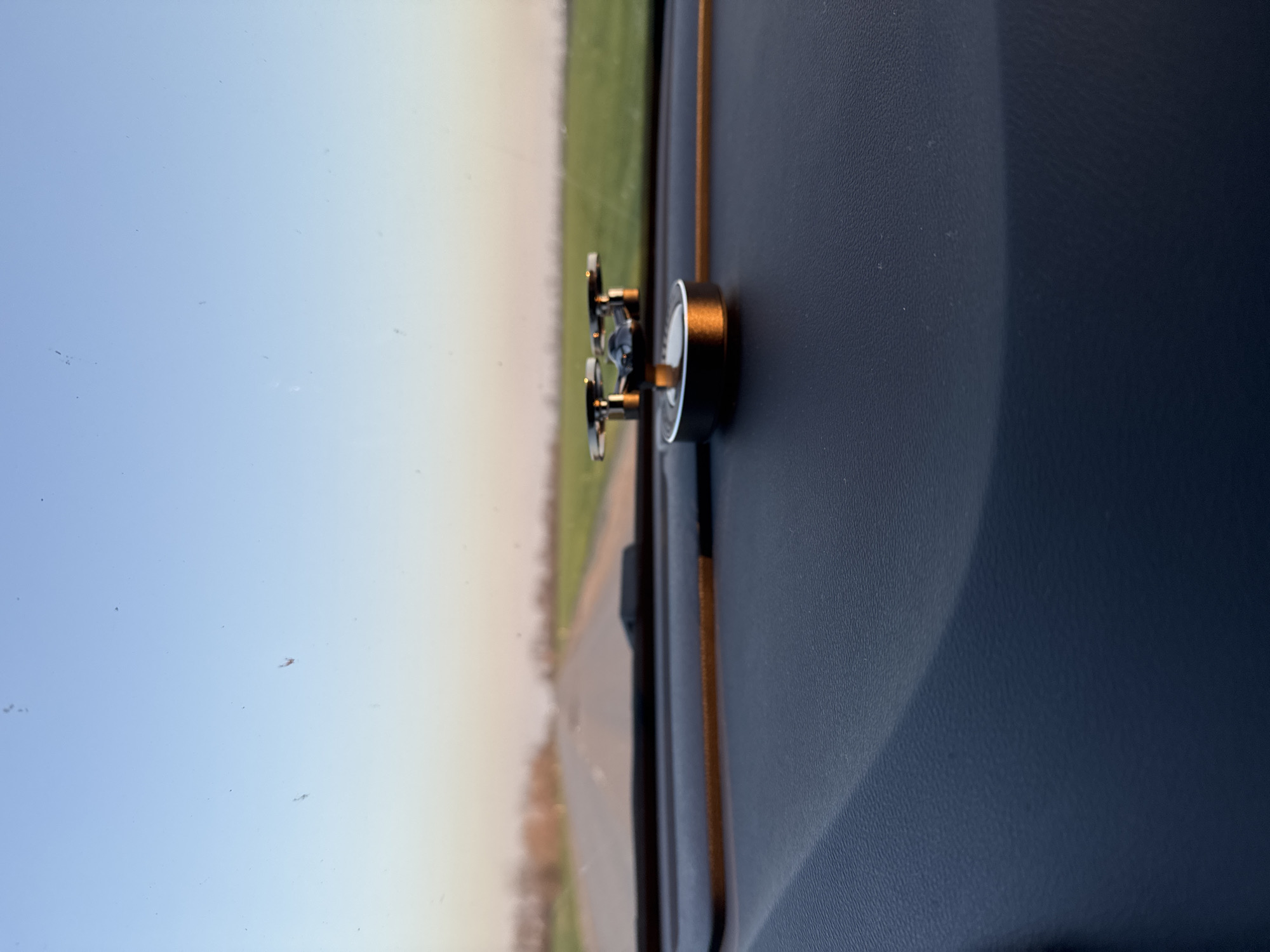 The image shows an analog gauge, likely a thermometer or pressure gauge, mounted on the dashboard of a vehicle. The gauge has a round face with a black bezel and is positioned close to the windshield. Outside the windshield, there is a view of a road and some green vegetation, suggesting a rural or suburban setting. The dashboard appears to be dark-colored, possibly black or dark blue. The image provides no information to determine the specific city or location.