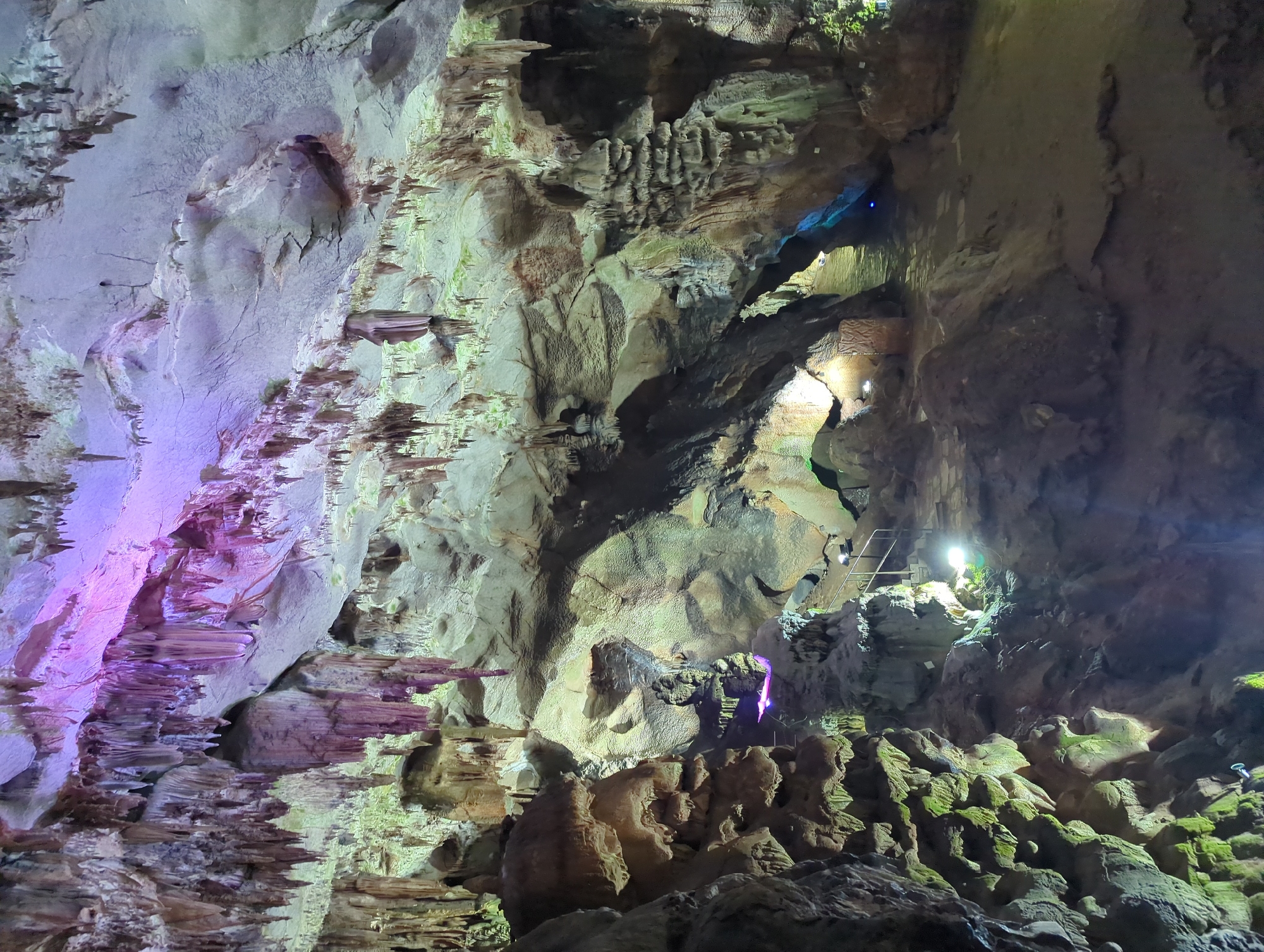 The image depicts the interior of a cave, likely a tourist attraction, with prominent stalactites and stalagmites illuminated by artificial lighting. The cave walls and ceiling are irregular and textured, showcasing natural rock formations. Several rock formations are highlighted with colored lights, including purple and green, creating a dramatic and surreal atmosphere. In the mid-ground, a set of stairs with handrails is visible, suggesting a pathway for visitors. The floor of the cave appears to be made of stone or compacted earth, with some areas appearing wet, possibly indicating a water feature or damp conditions. This scene is characteristic of the Vratsa Cave, a natural landmark located in the Vratsa Mountains near the city of Vratsa, Bulgaria, known for its impressive speleothems and accessibility to tourists.