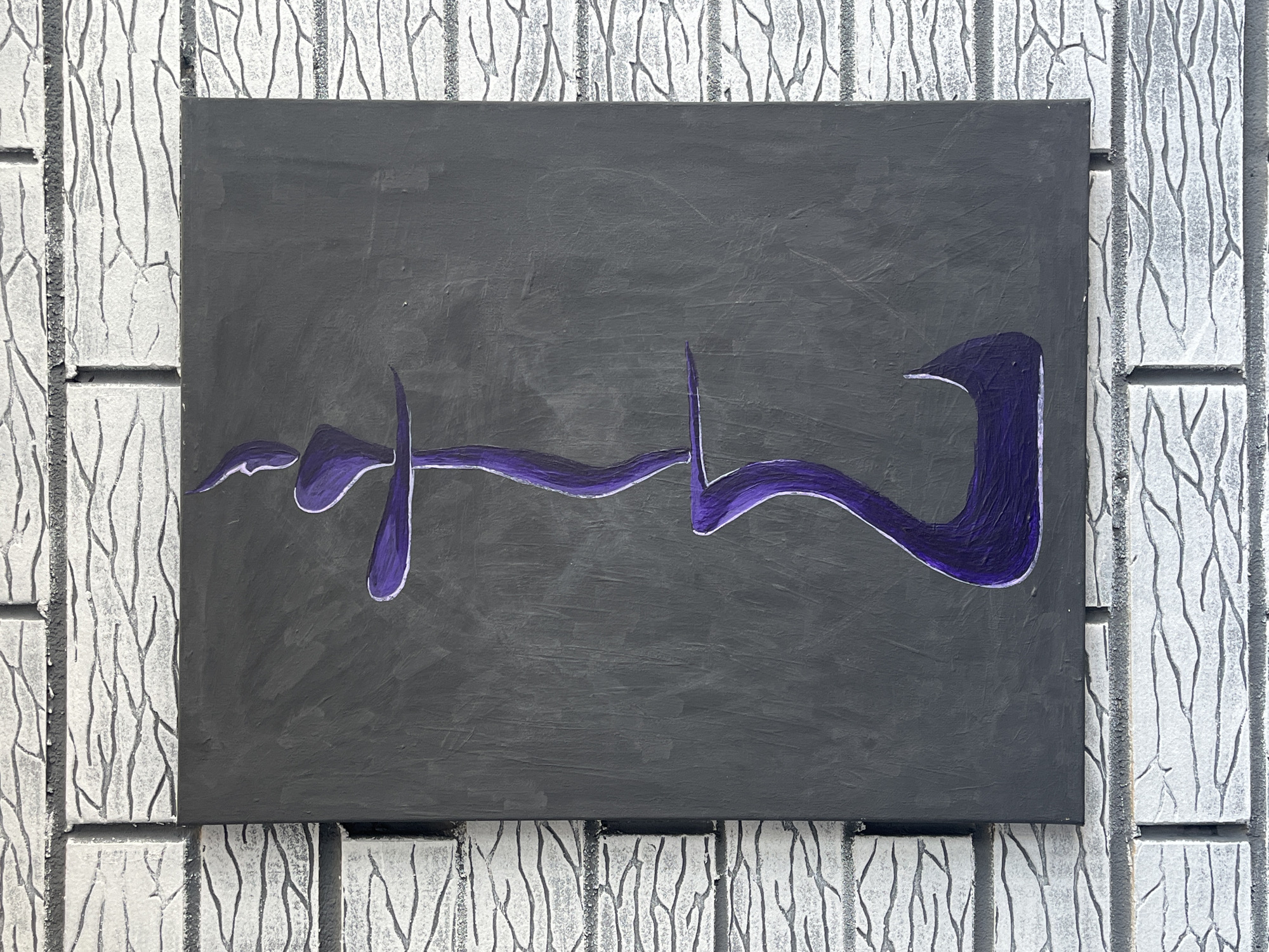 A vertical canvas painting with a dark gray background is displayed against a white brick wall. A stylized, abstract figure painted in purple with white outlines is centered on the canvas. The figure is composed of two main sections. The upper section is thinner and has a curved, elongated shape with a pointed top. It is balanced on a horizontal purple line. The lower section is wider and bulbous at the bottom, tapering upwards to connect to the horizontal line. The overall impression is of a flowing, organic form. The brick wall features a textured, irregular pattern in shades of white and gray, with dark lines indicating the mortar.