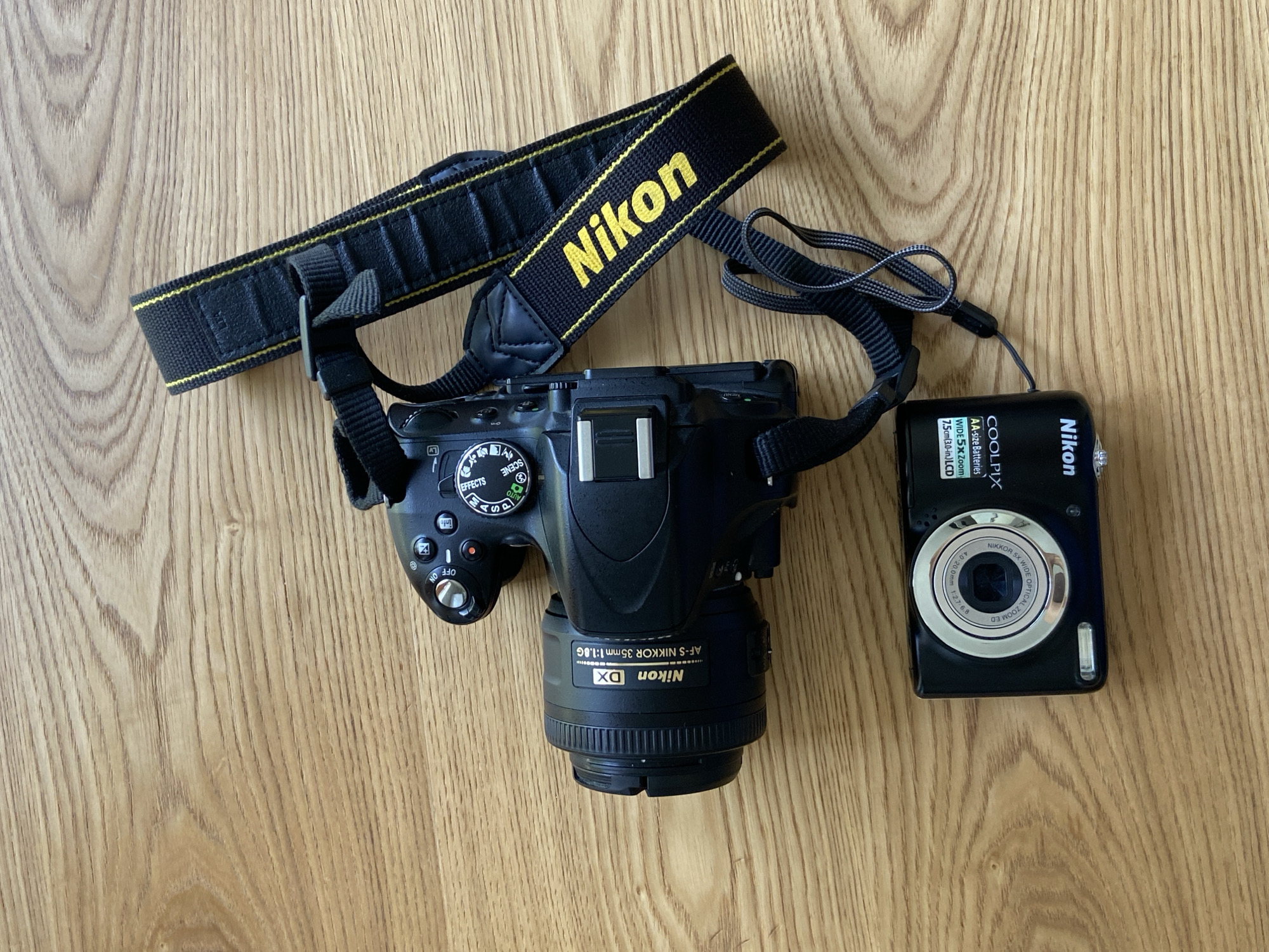 Two black Nikon cameras are displayed on a light-colored wooden surface with prominent grain, captured from an overhead perspective. On the left is a larger DSLR-style camera with a black and yellow "Nikon" branded strap. The lens attached to it is an "AF-S NIKKOR 35mm 1:1.8G Nikon DX". The camera's mode dial shows settings such as "AUTO", "P", "S", "A", "M", "EFFECTS", and "SCENE", and an "OFF ON" power switch is visible. To the right is a smaller, black compact digital camera. Its lens is labeled "NIKKOR 5X WIDE OPTICAL ZOOM ED 4.0-20.0 mm 1:2.7-6.8". Text on the camera body identifies it as a "Nikon COOLPIX" model, noting it uses "AA-size Batteries", has "WIDE 5x Zoom", and a "7.5cm (3.0-in.) LCD". A thin black wrist strap is attached to the compact camera. The objects are arranged statically, suggesting a flat-lay or product display. The scene is set in Tyumen, Russia.