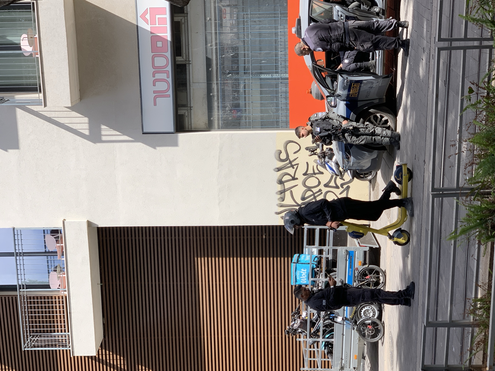 The image shows a street scene in Tel Aviv, Israel. On the right, two Israeli police officers are near a police car and a motorcycle, apparently in conversation. Further down the sidewalk, there is a Wolt delivery worker walking next to a trailer loaded with bicycles. Another person is on a lime green scooter. Behind the police officers is a white building with a business sign in Hebrew. Behind the delivery worker is a building with a dark brown vertical striped facade.