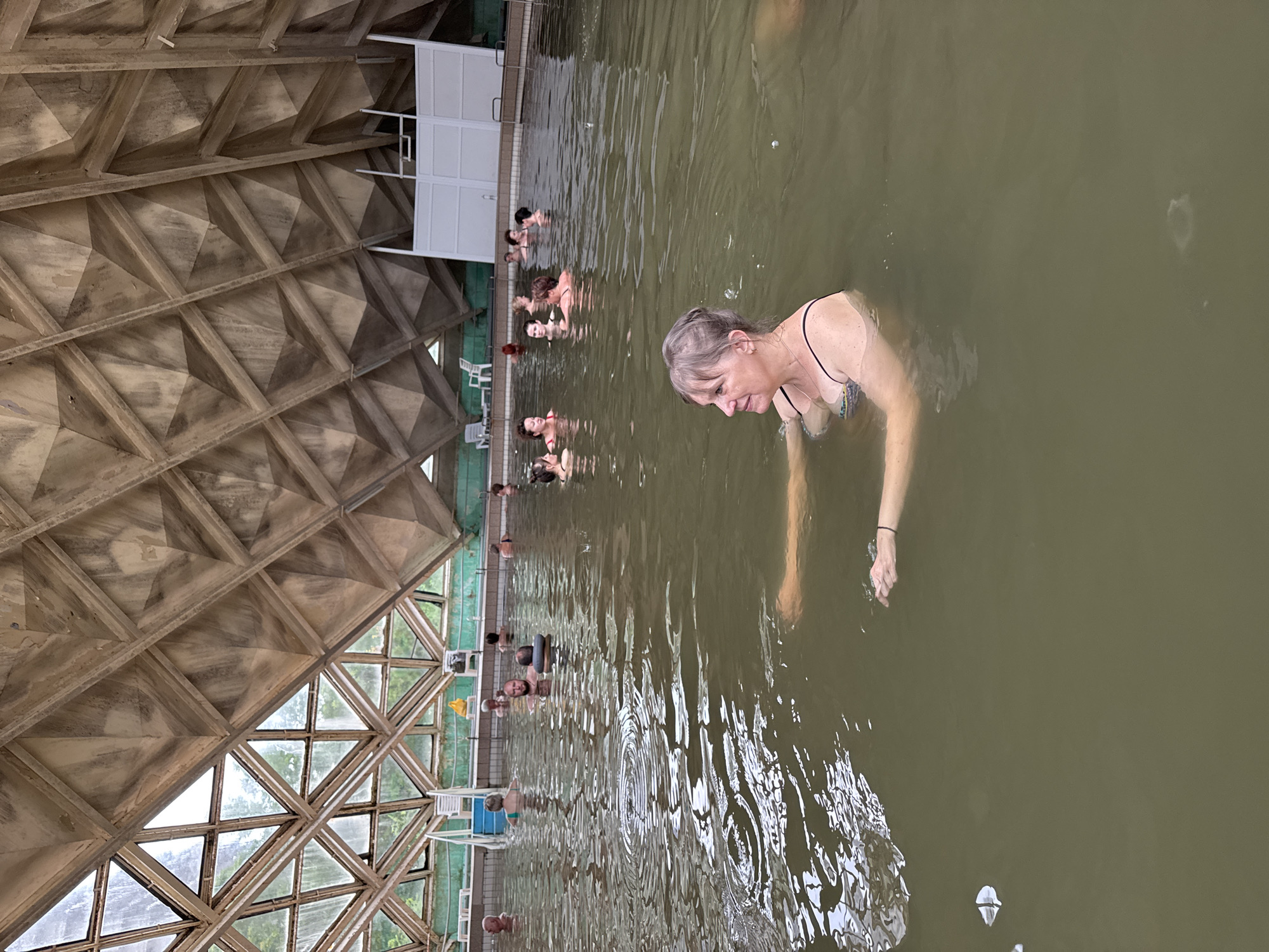 The image depicts an indoor swimming pool with a distinctive geodesic dome-like ceiling. Several people are visible in the water, most of whom are women wearing swimsuits. One woman in the foreground, with graying hair, is looking down and appears to be in the process of moving through the water. In the background, a line of people are standing or sitting in the pool, some with their heads visible above the water. The pool water is a murky greenish-brown color.
To the left of the pool, white structures resembling changing rooms or shower facilities are visible. Beyond the pool's edge, there are windows that suggest an outdoor area with greenery, indicating it might be daytime. The architectural style of the ceiling, with its repeating geometric patterns and exposed concrete, suggests a mid-20th-century or Brutalist design. The overall atmosphere seems relaxed, with people enjoying the pool. No specific time of day or weather conditions can be definitively determined, other than the presence of natural light from the windows. The location is stated to be Odesa, Ukraine. There is no visible text in the image.