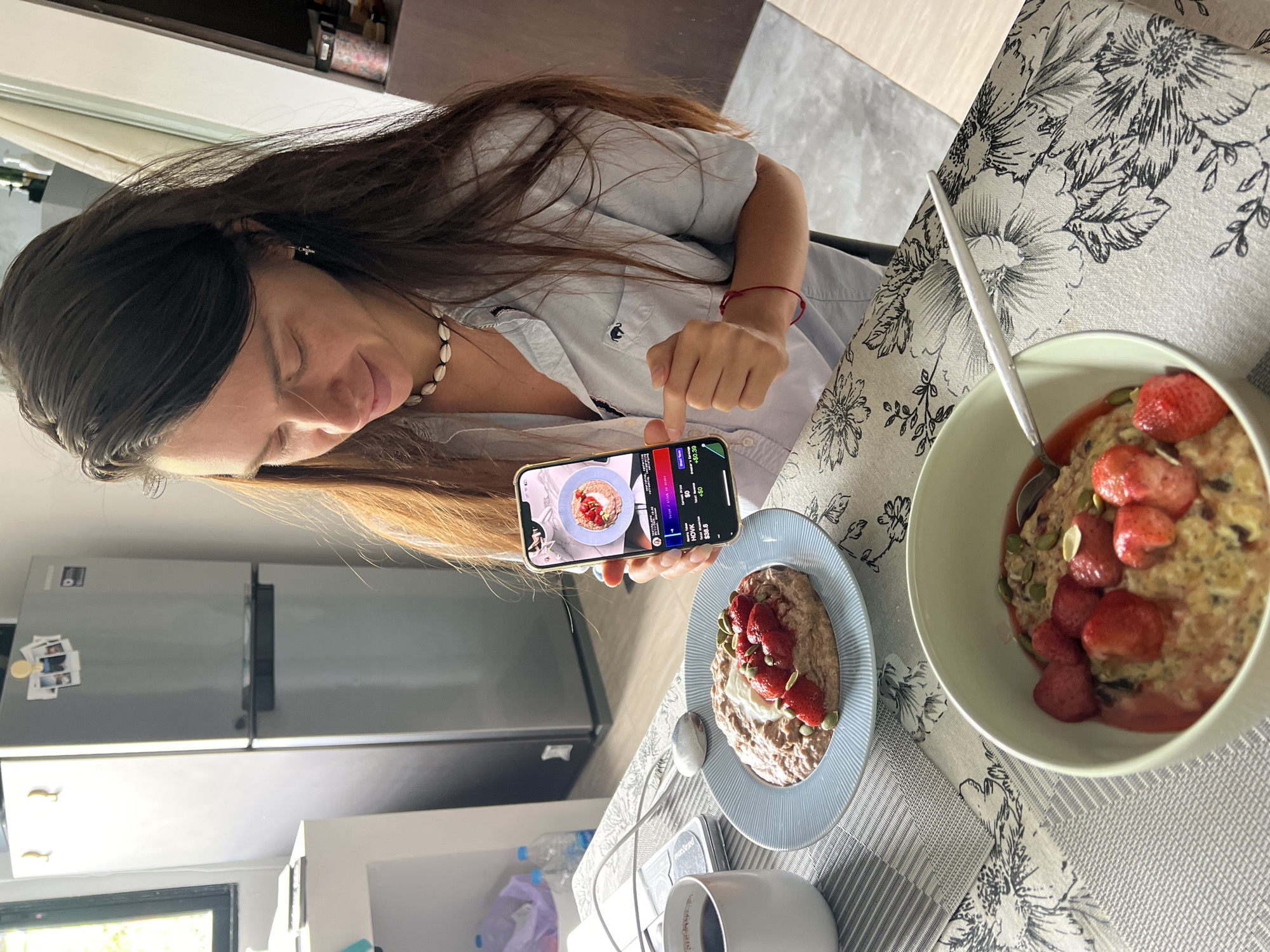 A woman with long dark hair, wearing a light blue collared shirt and a shell necklace, is seated at a table in what appears to be a kitchen. She is holding a smartphone in her right hand, displaying a graphic interface with financial data. Her left index finger is extended, possibly interacting with the screen.
On the table in front of her are two bowls of oatmeal topped with strawberries and seeds. A white mug, likely containing coffee, is to her left. In the background, a silver refrigerator is visible, adorned with several small photographs. A window is also present to the left, letting in natural light. The table is covered with a patterned cloth depicting floral designs. The overall environment suggests a domestic setting in Baan Bang Rak, Thailand.