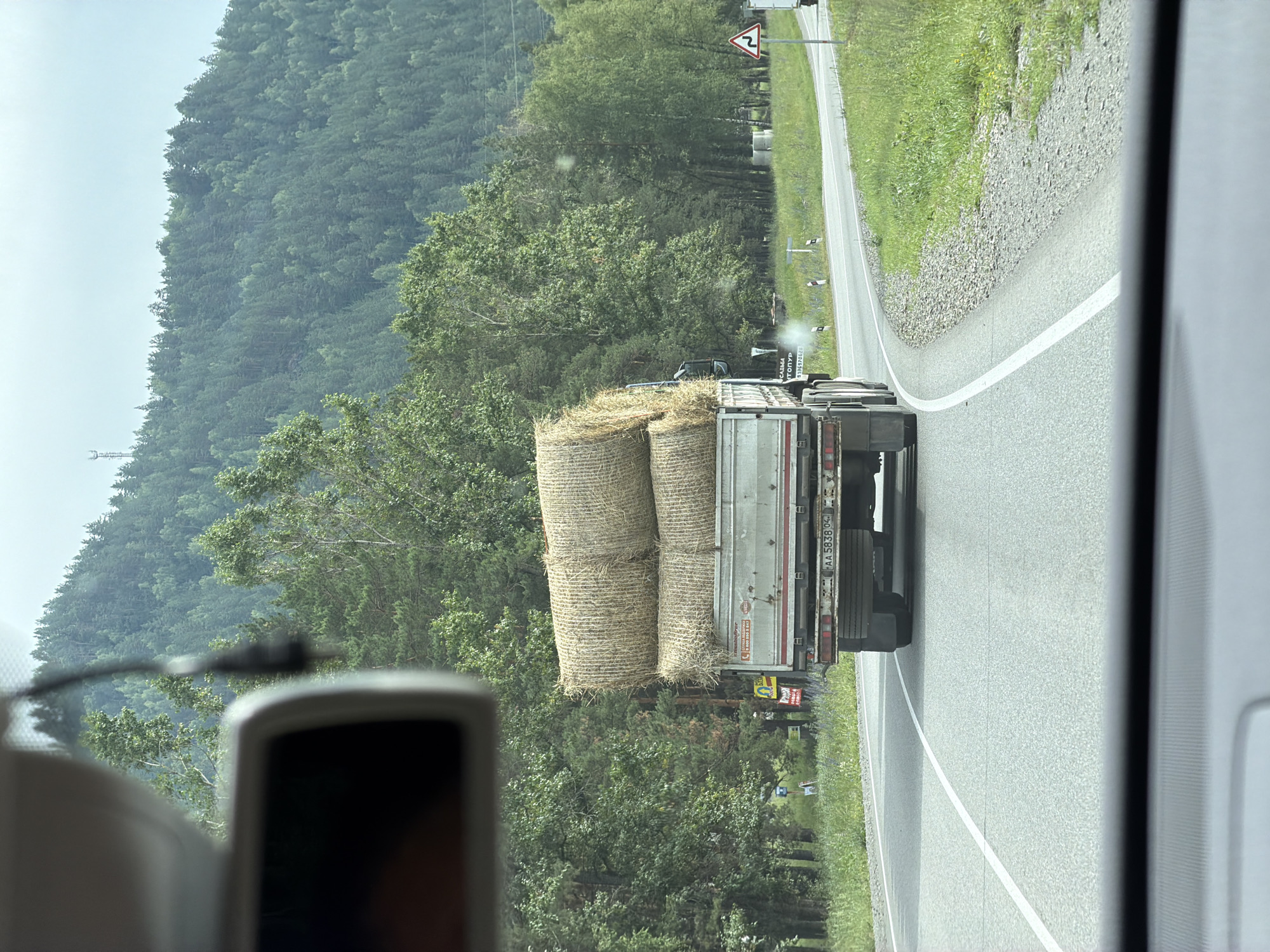 The image, taken from inside a vehicle on a cloudy day, captures a truck carrying a load of hay bales driving on a paved road. The truck is silver with a red accent on the back and has the license plate AA 5838 04. It is moving away from the camera on a winding road that passes through a rural, mountainous area. Dense green trees cover the slopes of the mountains in the background, and a radio tower is visible on one of the peaks. The foreground shows the dashboard and a portion of the windshield of the vehicle from which the photo was taken. The surrounding environment appears to be the area around Катунь, Russia, characterized by its natural landscape and a road designed for vehicles.