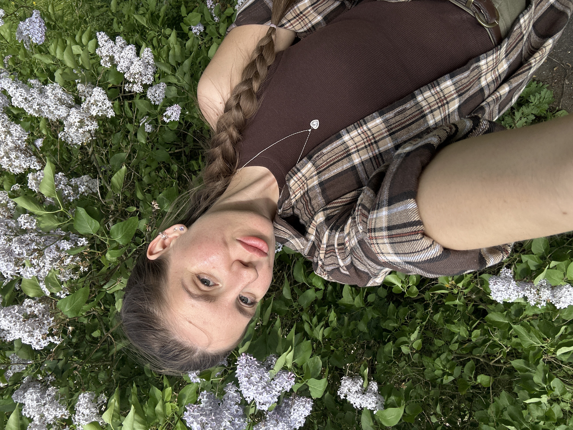 A young woman lies in a field of lilac flowers in Борщевка Малая, Russia. She has light skin, blue eyes, and her brown hair is braided. She wears a brown ribbed tank top with a silver heart-shaped necklace and a brown plaid flannel shirt.