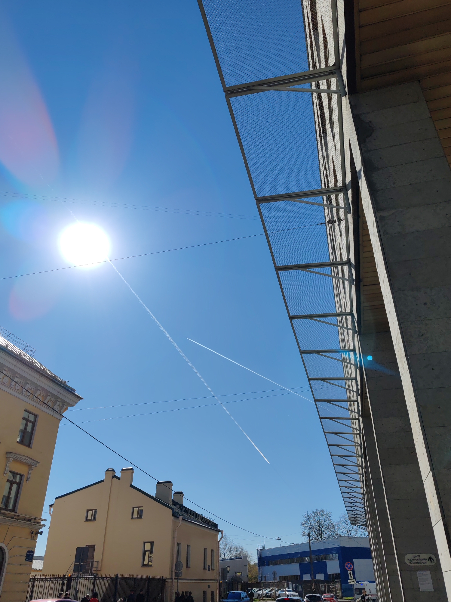 This vibrant image captures a sunny urban street scene in Saint Petersburg, Russia, under a clear blue sky. The bright sun, positioned in the upper left, creates a prominent lens flare, dominating the sky. Two distinct airplane contrails streak across the azure expanse, one long and leading directly from the sun, and another shorter one intersecting it.
In the immediate foreground on the right, a large, modern concrete structure with a grid-like metal mesh attachment extends vertically, suggesting part of an elevated walkway or building facade. Visible on a sign attached to this structure is Russian text: "ЦЕНТР ИЗГОТОВЛЕНИЯ ИЗДЕЛИЙ ВСЕ" (CENTER OF MANUFACTURING PRODUCTS ALL) accompanied by a right-pointing arrow.
On the left, an ornate, light-colored historic building with dark windows stands prominently, contrasting with a simpler, cream-colored building featuring multiple chimneys and dark windows further back. Several overhead power lines crisscross the scene.
The mid-ground reveals a bustling street with various cars parked and moving, including an ambulance in the distance. Several pedestrians are visible on the sidewalk. In the background, a distinctive blue and white industrial-style building can be seen. Bare trees, hinting at early spring or late autumn, are scattered throughout the scene. A traffic sign with a red circle and white bar (likely "No Stopping" or "No Entry") is also visible.