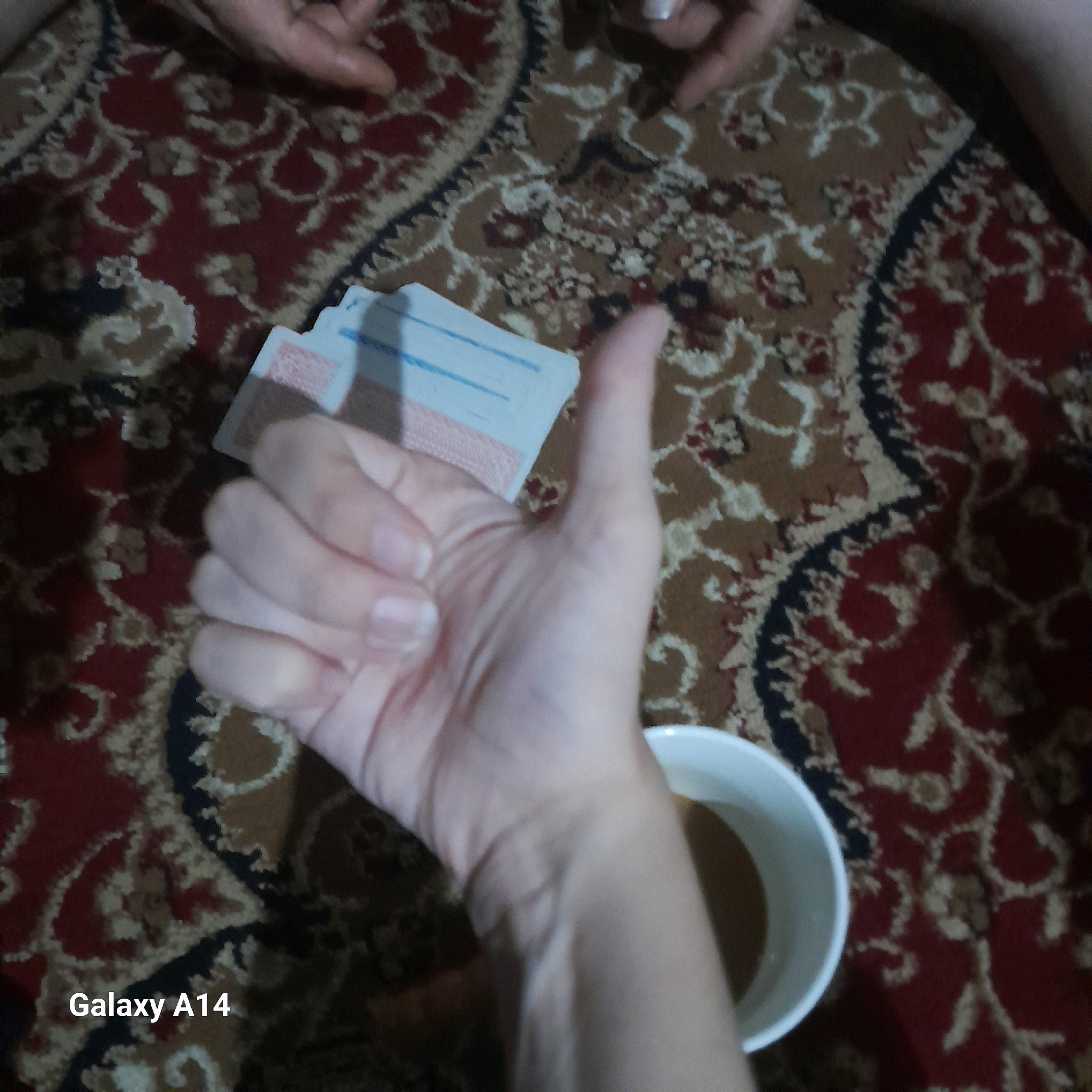 The image, watermarked with "Galaxy A14", is an overhead shot of a patterned rug in what appears to be a residential setting. A hand with manicured fingernails is prominently featured, giving a thumbs-up gesture. To the left of the hand, a small stack of what looks like playing cards or similar small rectangular objects with blue and red markings is visible. To the right of the hand, the edge of a white mug containing a brown liquid, likely coffee or tea, is present. The background is a dark, intricately patterned rug with red, brown, and tan elements. The image does not provide any specific visual information that would allow for its location to be determined as Khan Yunis, Palestinian Territories.