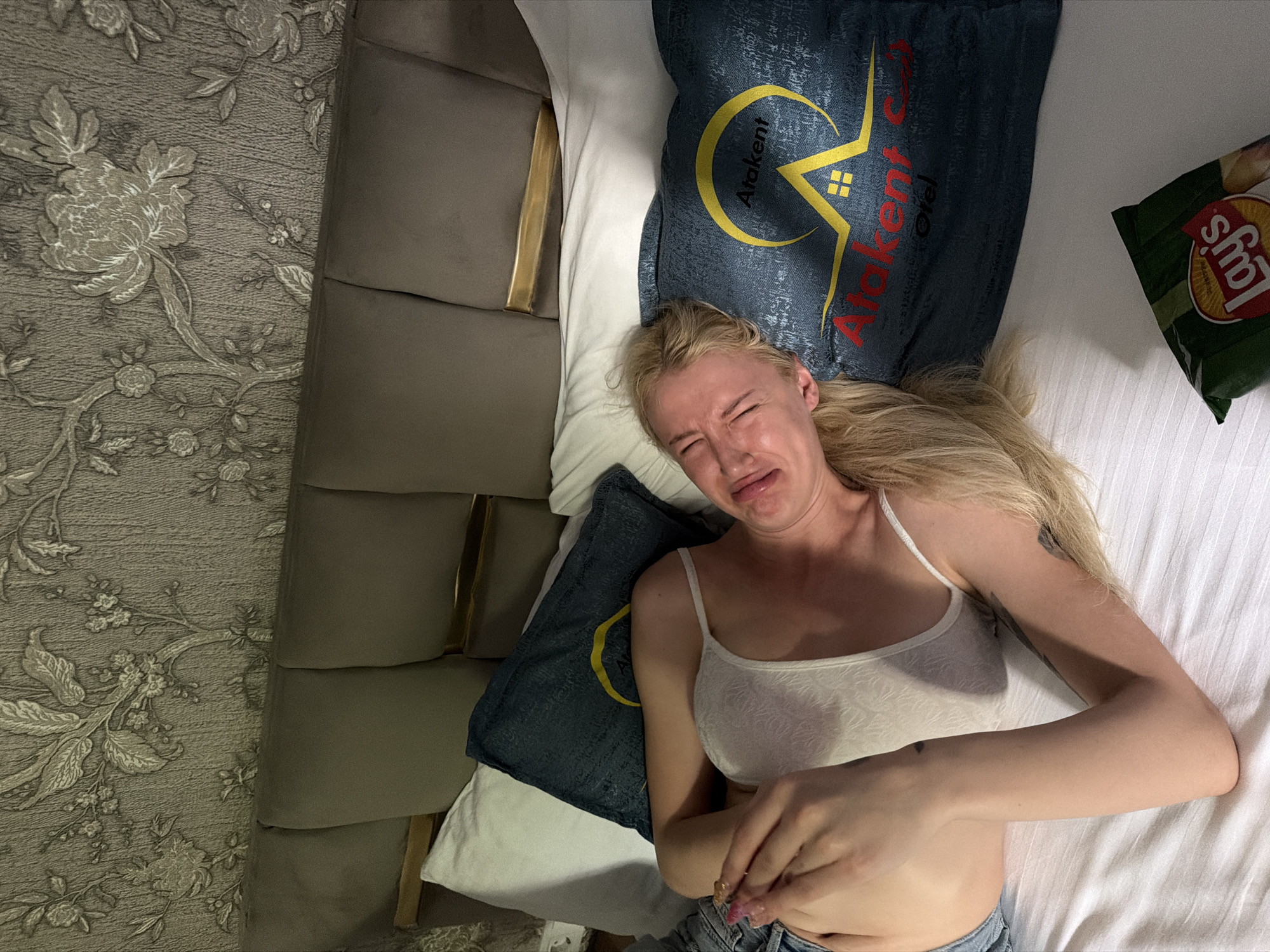 A woman with blond hair lies on a bed in what appears to be a hotel room. She is wearing a white lace top and denim jeans. Her face expresses distress, as if crying or in pain. Pillows with the logo of "Atakent Otel" are visible near her head. A bag of Lay's potato chips rests near her arm. The location can be inferred to be in Atakent, Küçükçekmece, Turkey, based on the hotel logo on the pillows. The scene is captured from an overhead perspective.