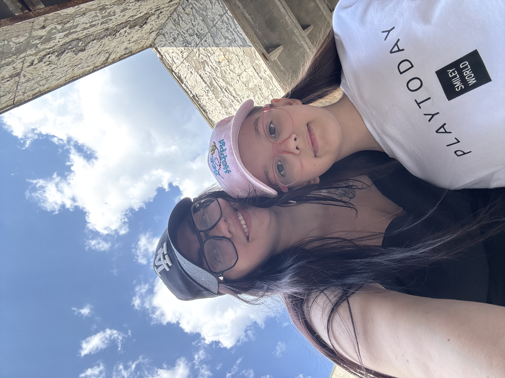 Two individuals are visible in the image, positioned closely together, suggesting a selfie. One individual, presumably an adult, is wearing a dark baseball cap with a white logo (potentially "NY") and glasses. The other individual, likely a child, wears a pink cap with a design and pink-rimmed glasses. The child is wearing a white t-shirt with the words "PLAY TODAY" and the phrase "SMILEY WORLD" on it. They are positioned against a backdrop of a blue sky with scattered clouds. Above them is a structure made of stone. The location is Unknown.
