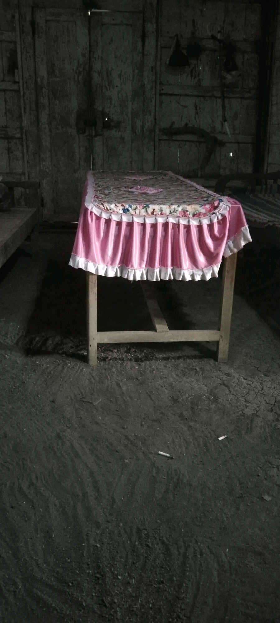 A table draped with a pink tablecloth with a floral pattern and white trim stands in a dimly lit room. The room appears to be made of wood and has a dirt floor. There are two cigarette butts on the floor near the table. The overall impression is one of neglect and disuse.