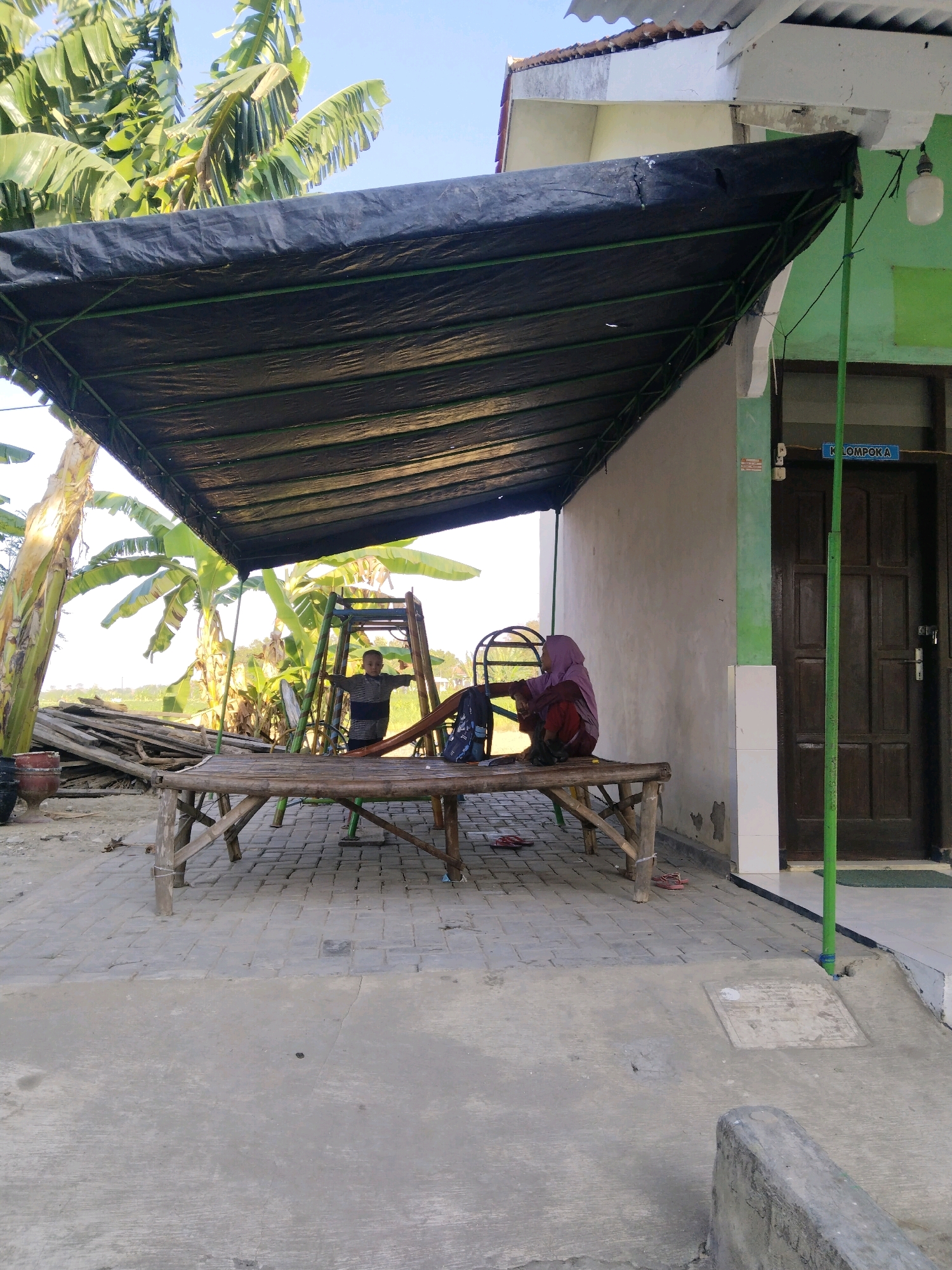 A medium shot captures an outdoor scene in Kaduara Barat, Indonesia, under a large black tarp supported by a green metal frame. In the foreground, a rustic bamboo table with a bamboo bench is set up on a paved area. A person wearing a purple hijab and dark clothing sits on the bench, facing away from the camera and looking towards a building. To the left of the table, a child in a grey and black striped shirt stands on a small wooden playground structure. Banana trees with large green leaves are visible in the background, along with a stack of wooden planks. The building behind the tarp has a wooden door and a sign above it that reads "KELOMPOK A". The overall atmosphere is casual and appears to be a public outdoor space.