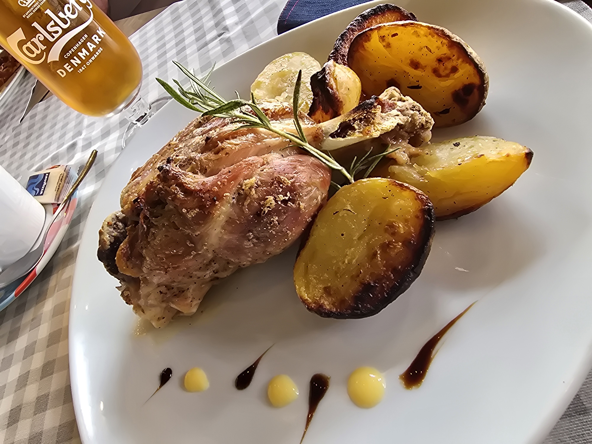 A close-up, overhead shot shows a white plate with a meal of roasted meat and potatoes. The meat, a large piece of pork, is seasoned and has a golden-brown crust. It is garnished with a sprig of rosemary. Several thick slices of roasted potatoes, some with charred edges, are arranged around the meat. Drizzled on the plate are small dollops of a yellow sauce and dark brown sauce. In the background, a checkered tablecloth and a glass bottle of Carlsberg beer are visible. The scene appears to be set in a restaurant or dining area.