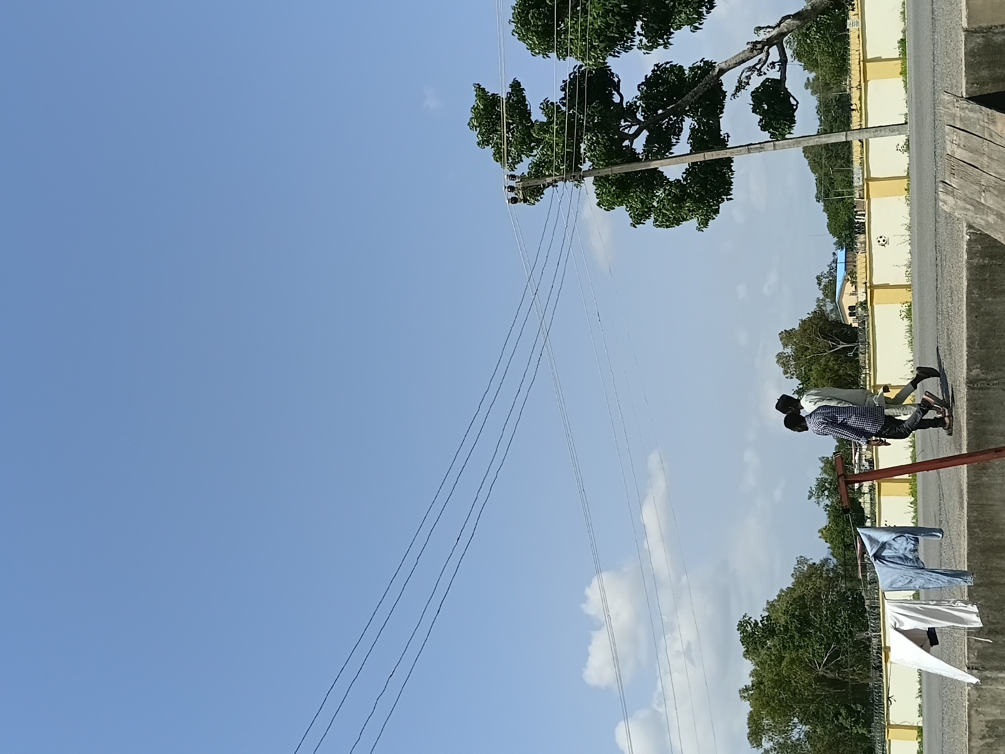 The image depicts a scene outdoors in Jalingo, Nigeria, under a clear blue sky with scattered clouds, suggesting daytime. A power line runs diagonally across the frame. In the foreground, a concrete barrier is visible, with clothing hanging on a line. Two young men are standing on the barrier, appearing to be conversing. One is wearing a blue checkered shirt and dark pants, and the other is wearing a white shirt and dark pants. Behind them, a yellow wall runs along a road, with trees and buildings in the distance. The overall atmosphere appears casual and unhurried.