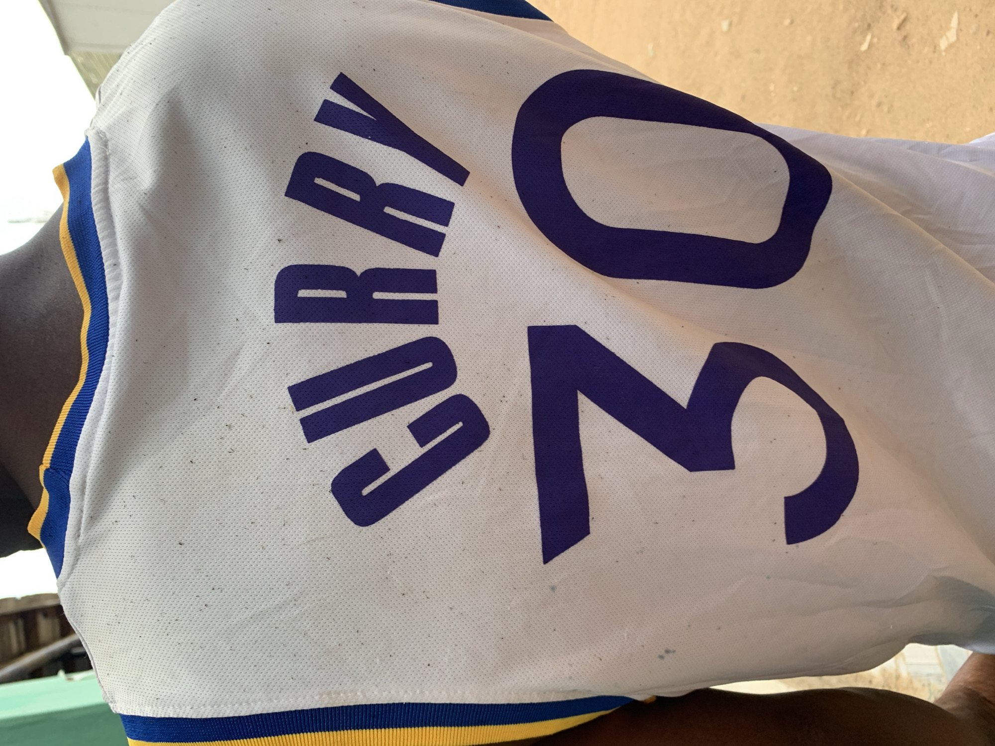 The image shows a person wearing a Golden State Warriors basketball jersey with "CURRY 30" printed in purple lettering on the back. The jersey is white with yellow and blue trim at the neck and armholes. The person's skin tone is dark. The setting appears to be outdoors. The city of New Stadium, Nigeria, cannot be confirmed from the provided image.