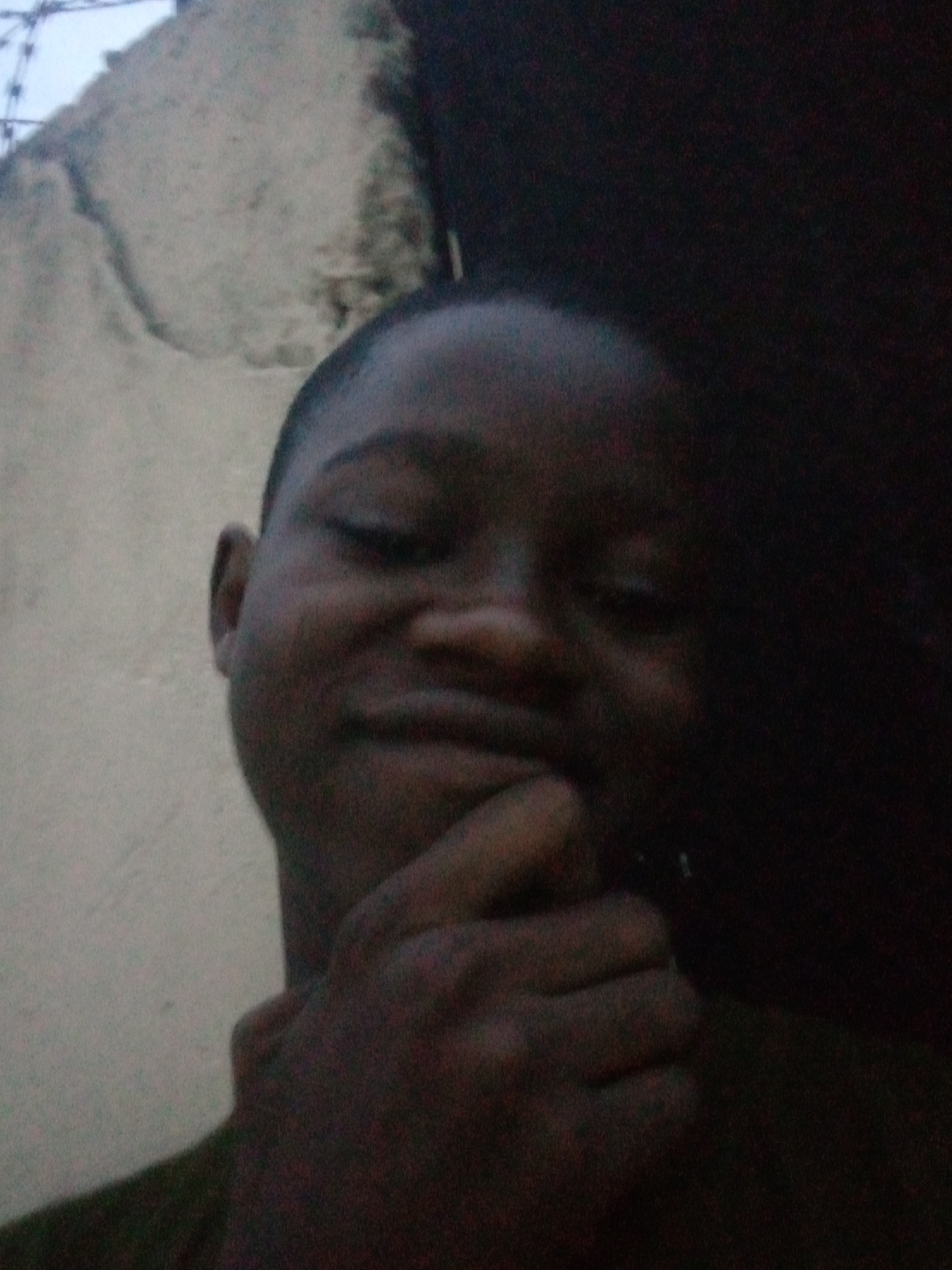 A low-light, slightly upward-angled close-up image depicts a young individual of dark complexion, situated in Shangisha, Nigeria. The subject's head and upper torso are visible, featuring short, dark hair. A faint, closed-mouth smile is present, with the eyes appearing partially obscured or lowered. The subject's right hand (viewer's left) is raised, with fingertips resting against the chin in a contemplative or posed gesture. The background on the left side reveals a light-colored, textured wall, possibly concrete or stucco, exhibiting irregular markings and a faint suggestion of a fence or wire structure at the upper left edge. The right side of the background is uniformly dark, indicating a deeply shadowed or unilluminated area. The image quality is grainy due to the dim lighting conditions.