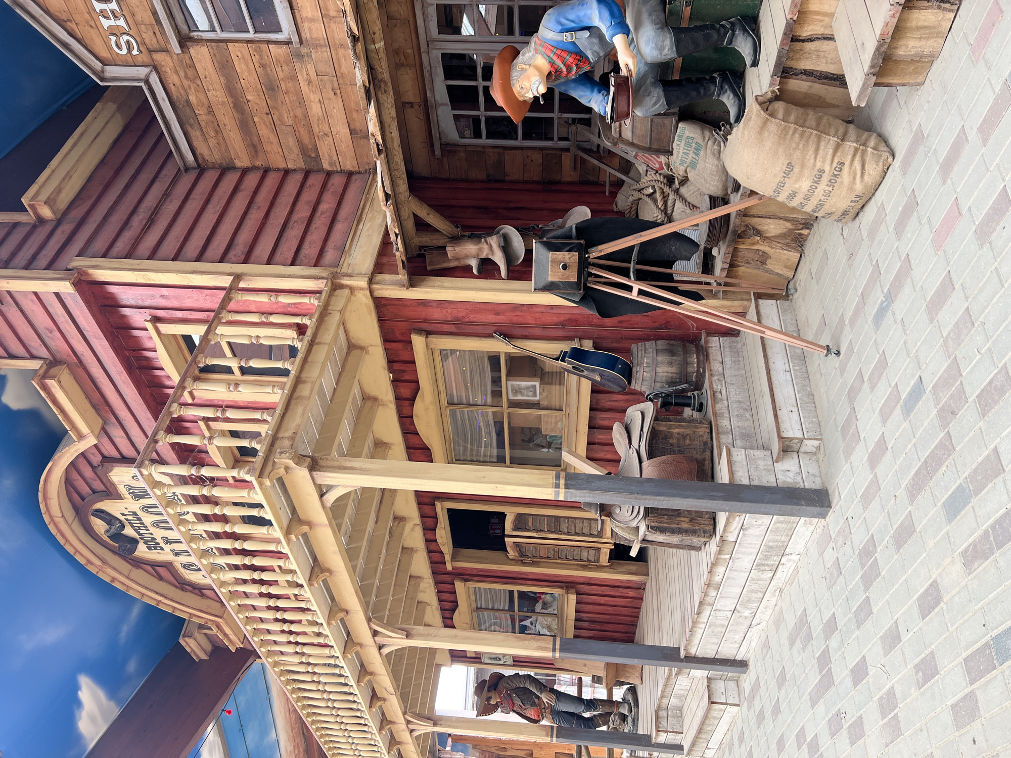 The image depicts a western-themed attraction, likely a restaurant or shop, in the city of Collendoorn, Netherlands. In the foreground, a paved area with a pattern of gray and reddish-brown bricks leads to the entrance of the establishment. The building itself is constructed with weathered red and brown wood siding and features a prominent second-story balcony with a wooden railing.
On the left side of the entrance, a life-sized mannequin of a cowboy stands on the wooden porch, wearing a brown hat, jacket, and jeans. Beside him, a wooden bench is visible. The main entrance has double swinging doors with wooden shutters.
To the right of the entrance, a second mannequin depicts an elderly cowboy seated on a wooden step. He wears a brown hat, a plaid scarf, and blue overalls, and holds a bowl and a cup. In front of him, there are several burlap sacks, one of which is labeled "COFFEE-14/LIP" and "WEIGHT: 50 KGS". Various other western-themed props are scattered around, including boots hanging from a beam, a vintage camera on a tripod, a saddle on a wooden crate, and barrels. A blue acoustic guitar leans against a barrel.
The facade of the building above the balcony displays a sign that reads "BOOTHLILL SALOON". The sky above is a bright blue with white clouds, appearing to be artificial, suggesting an indoor or partially covered space designed to simulate an outdoor environment. The overall scene is a static, staged display designed to evoke a Wild West atmosphere.