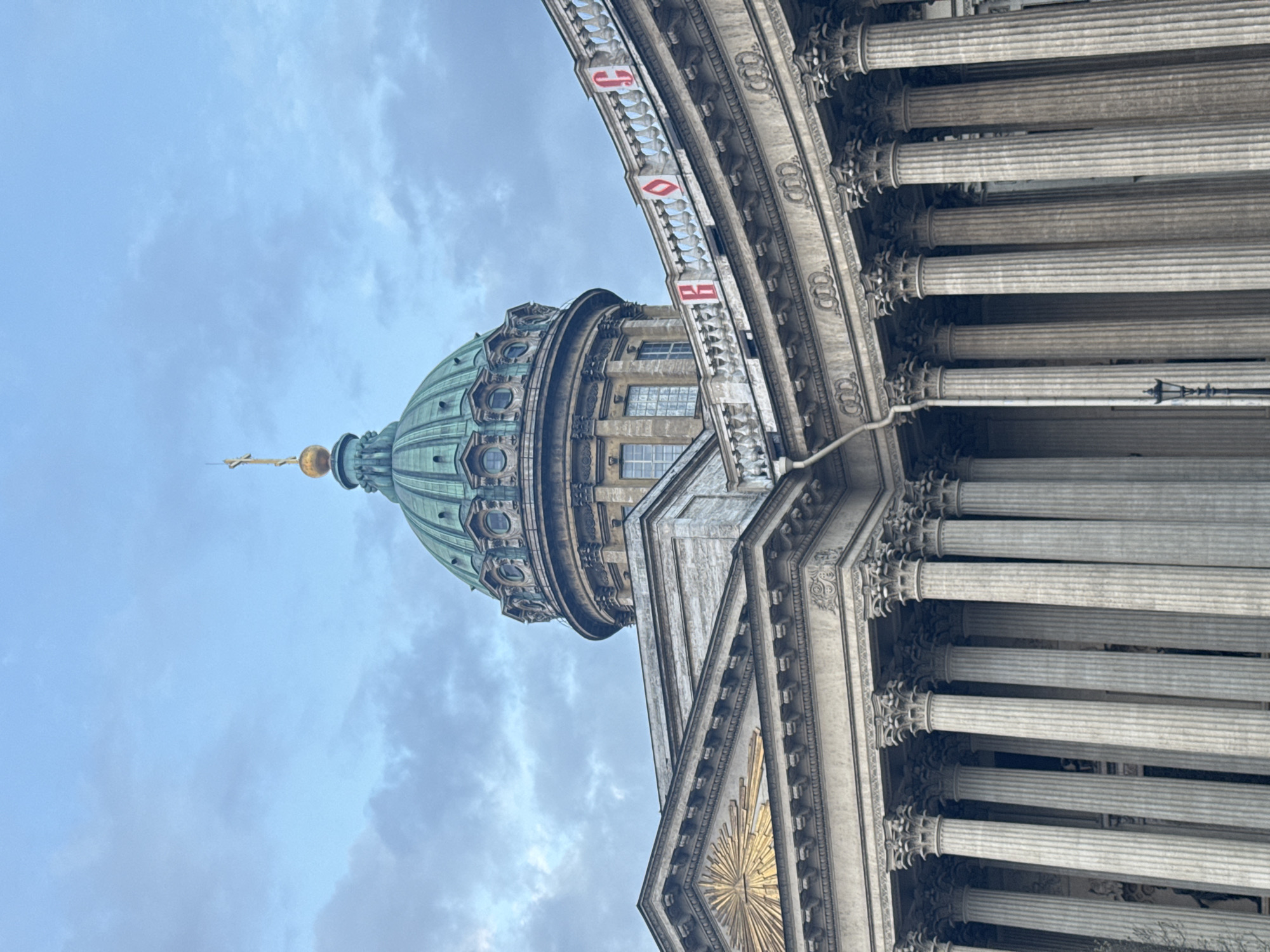 The image, rotated 90 degrees clockwise, presents a detailed view of a grand architectural structure, identifiable as the Kazan Cathedral in Saint Petersburg, Russia. The composition is dominated by a large, fluted green dome crowned with a golden finial and cross, soaring against an overcast, cloudy sky.
Below the main dome, intricate details of its drum are visible, featuring small windows and ornate decorative elements. To the right, a significant portion of a majestic colonnade is shown, comprising numerous tall, fluted columns with elaborate capitals, characteristic of neoclassical architecture. These columns are made of light grey stone, contributing to the building's imposing presence.
Between the dome and the colonnade, a curved stone balustrade is prominent, adorned with distinct red symbols on white rectangular panels. These symbols appear to be stylized Cyrillic letters, specifically "О" (or a similar circular/heart shape) and "Б" (or "B"), adding a unique decorative touch. Further down on the building's façade, a radiant golden sunburst motif is visible, emphasizing its grandeur.
The scene depicts no activity or people, focusing entirely on the monumental architecture. The overcast sky suggests a daytime setting with soft, diffused lighting, enhancing the texture and details of the stone and metalwork. The overall impression conveyed is one of historical significance and architectural splendor.