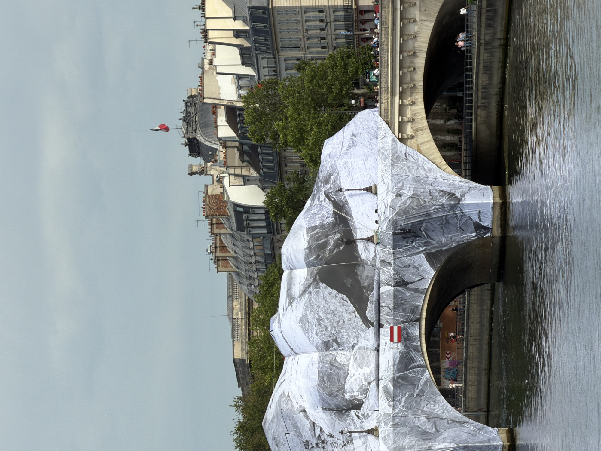 This is a photograph of the Pont Neuf bridge in Paris, France, as seen from the Seine River. The bridge is partially covered in a large, white fabric with a photographic print of a rocky landscape. Two arches of the bridge are visible, with the one on the left mostly obscured by the fabric covering. The fabric appears to be draped over the structure, creating a mountainous, textured surface. On the arch of the bridge on the left, there is a small, rectangular red and white flag.
The background shows Parisian buildings with mansard roofs and windows, typical of the Haussmannian architecture of the city. Green trees are visible behind the bridge, and the sky is overcast. People can be seen walking on the bridge and along the riverbank in the distance, with some figures also visible on the parapet of the bridge. A second, smaller bridge with multiple arches is visible in the background to the right. The water of the Seine River reflects the overcast sky and the surrounding structures.