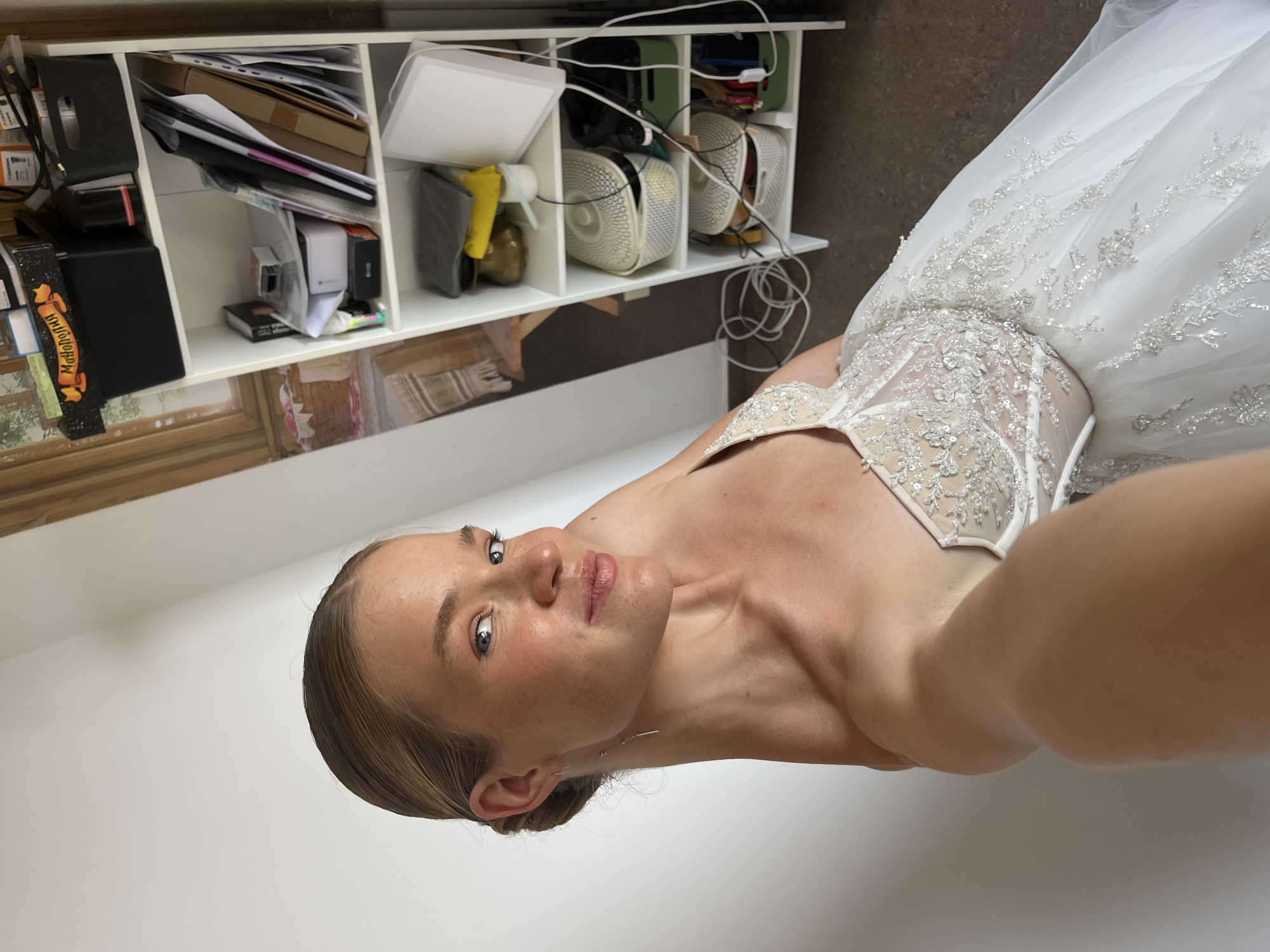 A young woman, viewed from a low angle, is in a close-up shot, wearing a white bridal gown adorned with intricate beading on the bodice and hem. The gown is detailed with delicate floral patterns and sparkling embellishments. The woman has her hair pulled back, and her face is turned to the side, looking upwards with a soft expression. She is wearing delicate earrings. In the background, a white shelving unit is visible, filled with various items including boxes, papers, and what appear to be personal belongings. The overall setting suggests an indoor environment, possibly a home or a bridal fitting room, given the presence of the wedding dress. The lighting appears to be natural, casting a soft glow on the subject. The image is oriented sideways.