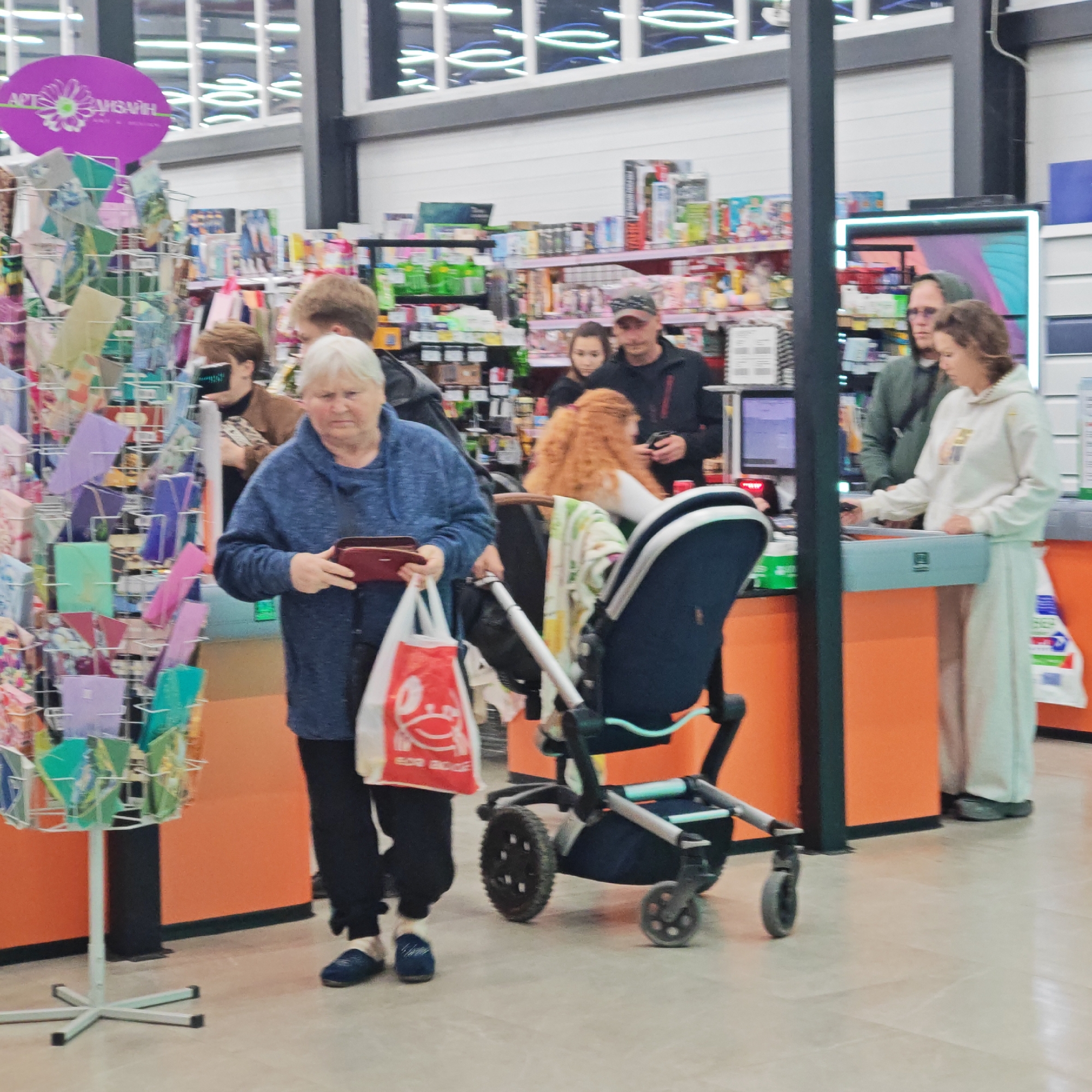 The image captures an indoor scene within a store, likely a supermarket or general merchandise outlet, focusing on a checkout area. Several people are present, engaged in shopping activities.
In the foreground, an elderly woman with white hair, wearing a blue sweater and dark pants, holds a red wallet in one hand and a white shopping bag with a red design in the other. The text "ВСЁ ДЛЯ ДОМА" (All for Home) is visible on the bag, along with a red smiley character. To her right, a dark-colored baby stroller with light blue accents and a patterned blanket draped over it stands next to her. Behind the stroller, a woman with long red hair is visible, and further back, a man in a dark jacket and baseball cap.
To the right side of the frame, a young woman in a white hooded sweatshirt and light-colored pants is standing at a checkout counter, seemingly making a purchase. A man in a green hooded jacket and glasses stands behind her. The checkout counters feature orange bases and light blue top panels, one of which displays a "回" symbol. Computer monitors and barcode scanners are visible on the counters.
The background shows numerous shelves stocked with various products, indicating a retail environment. A metal display rack with colorful greeting cards or small stationery items is positioned to the left. Above this display, a purple oval sign reads "АРТ СУВЕНІР" (Art Souvenir) with a daisy graphic, suggesting a gift or craft section. The store has a modern appearance with white walls, black structural pillars, and overhead fluorescent lighting that reflects in what appear to be high windows. The floor is covered with light-colored tiles. The Cyrillic text visible on signs aligns with the provided location context of Ukraine. The overall atmosphere is that of a busy but orderly retail environment.