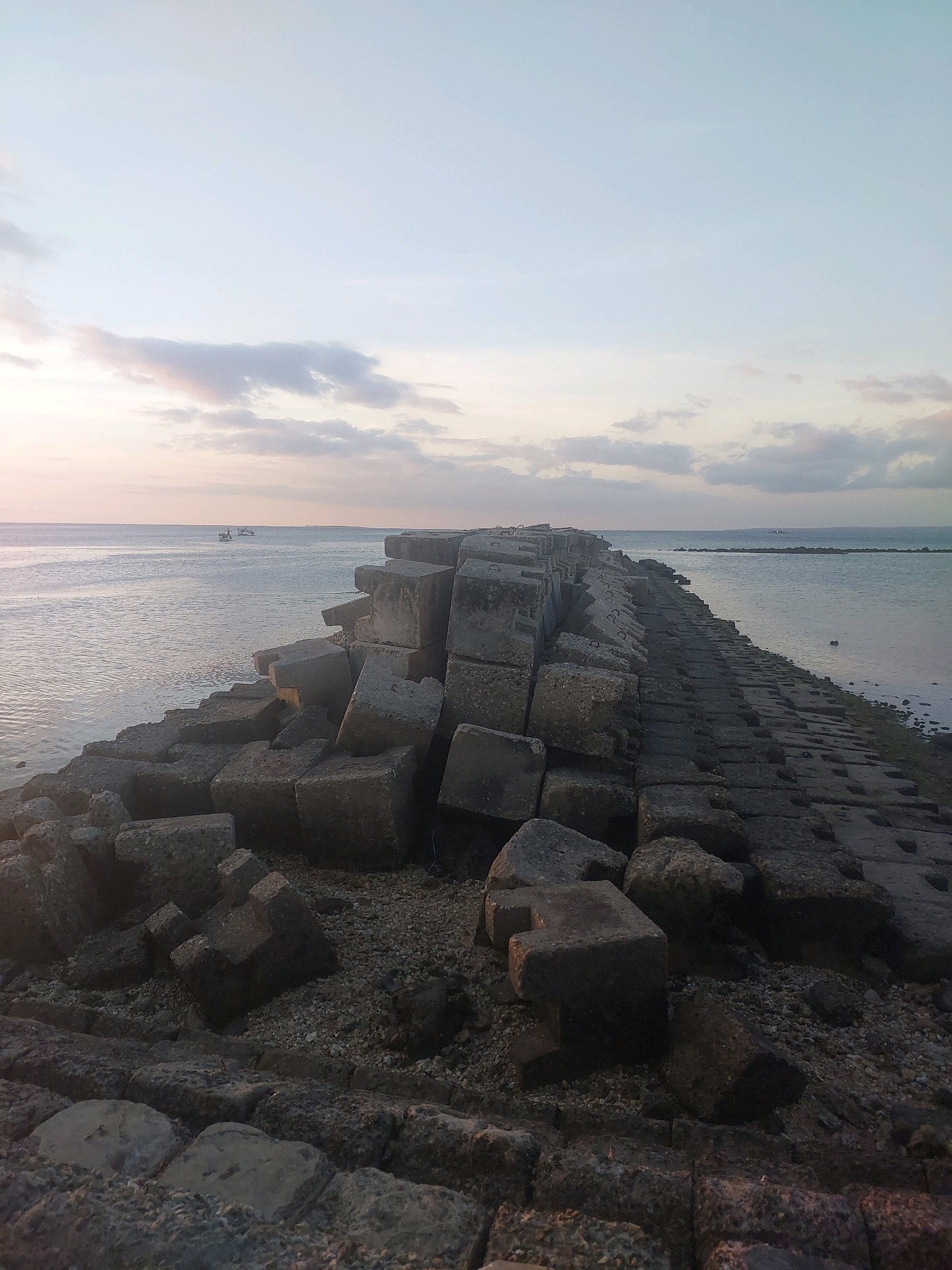 The image depicts a breakwater constructed from large concrete blocks extending into a calm body of water. The sky above is pale with scattered, wispy clouds, suggesting either dawn or dusk. In the distance, two small boats are visible on the water. The breakwater's surface is uneven, with many of the concrete blocks in the foreground appearing dislodged or precariously placed. The location is identified as the breakwater at Lasiana Beach in Kupang, Indonesia. The scene is devoid of any people, and no actions or interactions are observed. The environment is dominated by the man-made structure and the natural elements of the sea and sky.
