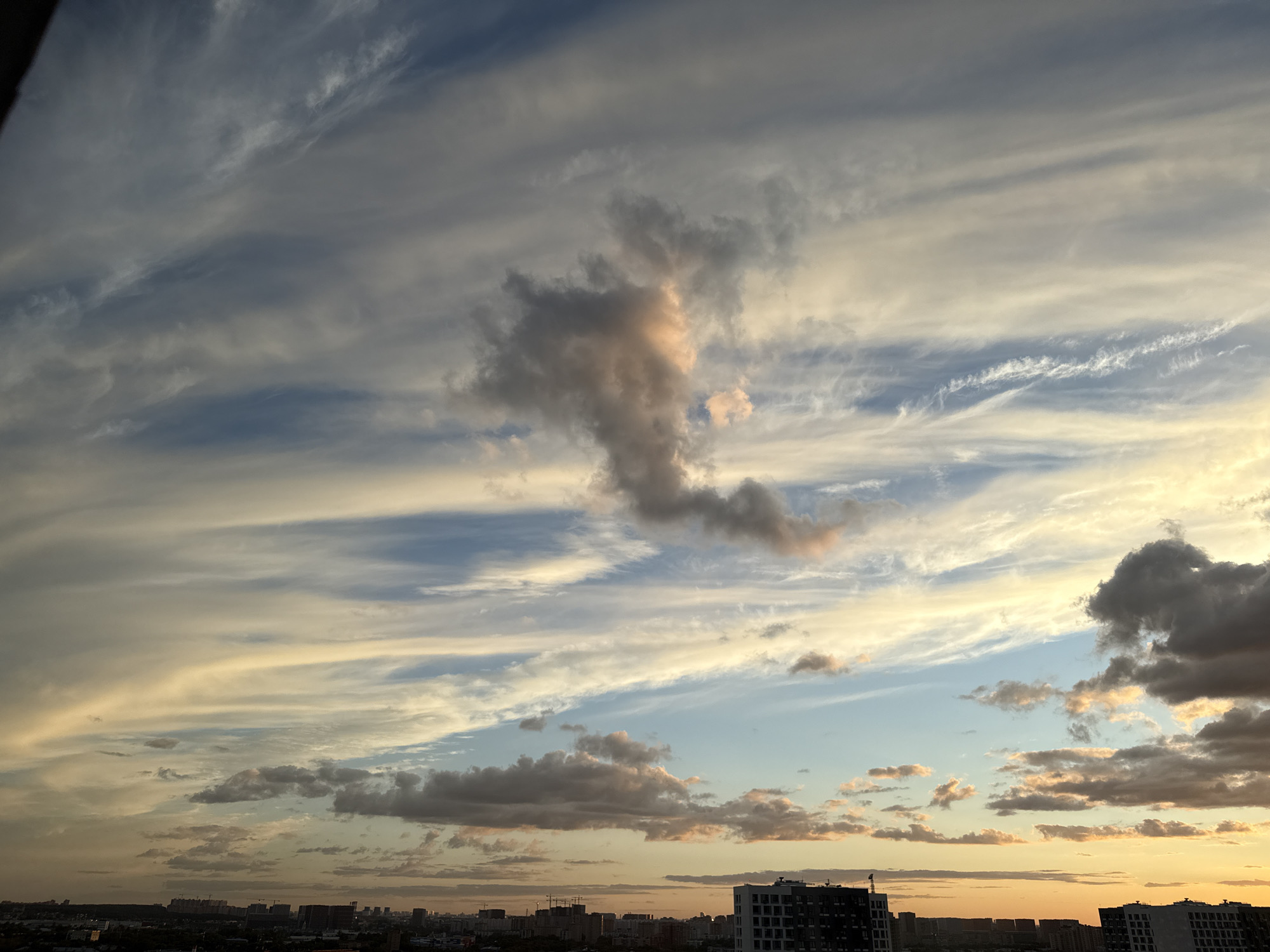 The image is a wide-angle shot of a cloudy sky over the city of Lyubertsy, Russia. The sun appears to be setting or rising, casting a warm, golden light on the clouds, with some areas appearing bright orange and pink. The clouds are varied in texture and form, with some appearing wispy and streaked, while others are more dense and textured, displaying shades of gray and white. In the lower portion of the image, the cityscape of Lyubertsy is visible, characterized by a collection of buildings, including apartment complexes and industrial structures. The buildings appear dark against the illuminated sky. There are no people or identifiable actions captured in the scene. The overall impression is one of a natural phenomenon occurring over an urban environment.