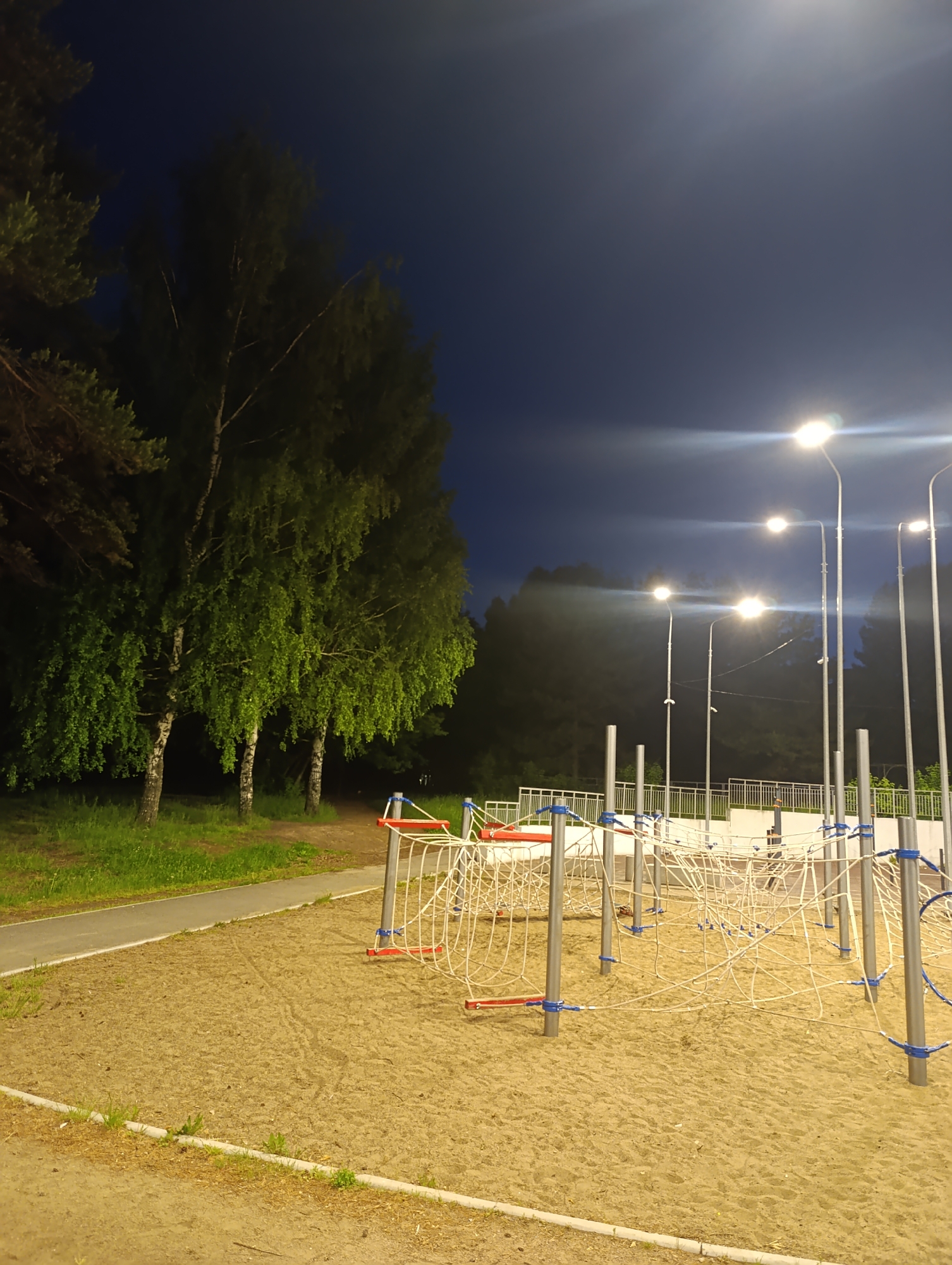 This nighttime photograph depicts a deserted outdoor playground. In the foreground and midground, a vast sandy area features an intricate climbing structure composed of white ropes woven around silver-colored poles, which are accented with red and blue elements.
To the left of the sandy playground, a paved pathway curves gently, bordered by a strip of green grass. Beyond this path, a dense line of tall trees stands, some exhibiting light-colored bark and lush green foliage, while darker, less defined trees extend into the background.
On the right side of the image, several tall streetlights illuminate the scene, casting bright glows with noticeable lens flares against a dark blue night sky. Behind the main playground equipment, a light-colored metal fence is visible in the midground. The sky above is clear, indicating fair weather. The overall setting suggests a public park or recreational area after dark. No people or visible text are present, conveying a quiet and peaceful, yet slightly vacant, atmosphere.