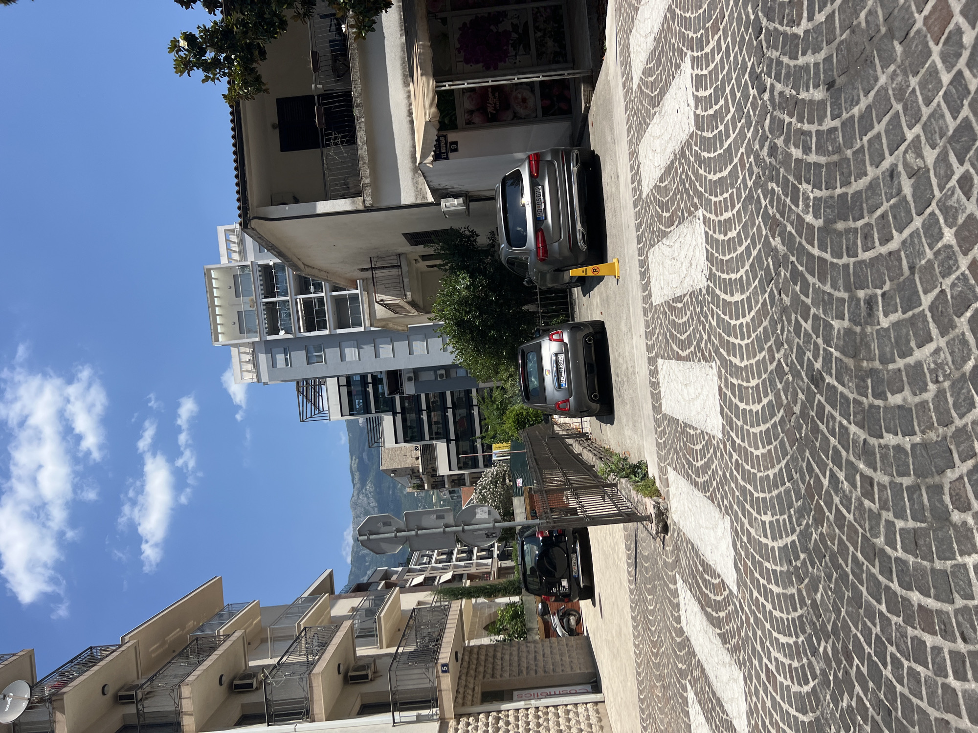 The image shows a street scene in Budva, Montenegro, on a sunny day. The perspective is from street level, looking up at buildings and down at the cobblestone street.
Dominating the foreground is a pedestrian crossing with white stripes on a patterned cobblestone surface. To the right of the crossing, a dark-colored car is parked. Further up the street, a silver car is also parked, with a yellow cone to its right.
The background features a mix of buildings, from older, lower-rise structures with balconies to a more modern, taller building with many windows. Some of these buildings have air conditioning units visible. Lush green trees are interspersed among the buildings. In the distance, a mountain range is visible under a bright blue sky with scattered white clouds.
There are no people visible in the image. The overall impression is one of a quiet, daytime scene in a European town, likely during warm weather given the clear sky and visible foliage.
Visible text includes license plates on the cars: "BD BS 872" on the silver car and "BD BB 270" on the black car. A small sign with the number "9" is visible on one of the buildings. Another sign with a red circle and a diagonal line within it, indicating a prohibition, is partially visible. One of the shop windows has the word "Cosmetics" printed on it.