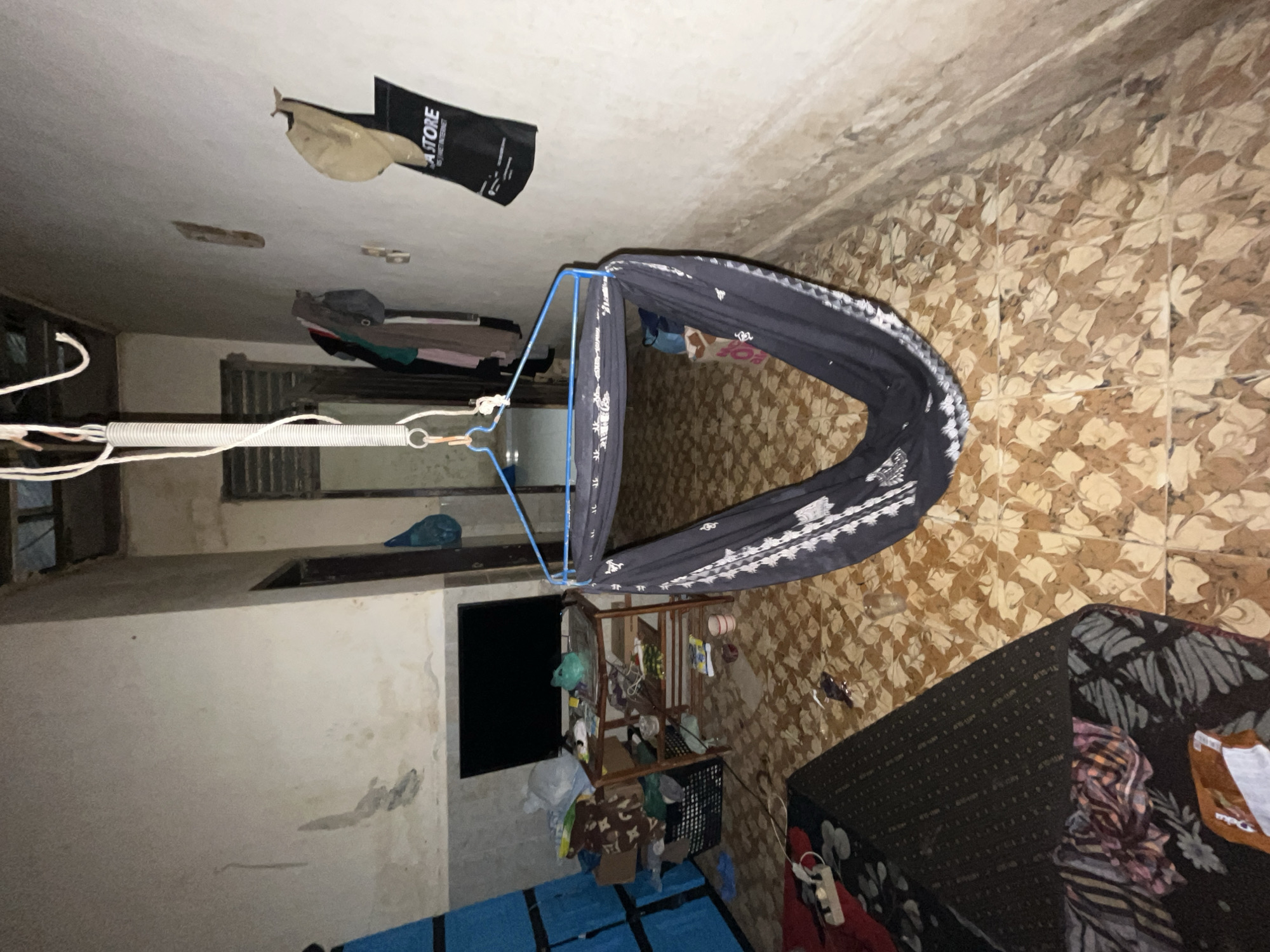 The image depicts an indoor scene, possibly a room or a hallway, with elements suggesting a living space in an Indonesian context, specifically Kota Kuala Simpang. The orientation of the image is unusual, rotated approximately 90 degrees clockwise.
The primary subject is a dark-colored hammock with a white patterned trim, suspended by ropes and a spring from what appears to be the ceiling. The hammock is empty. Nearby, a metal frame supports a smaller structure with various items hanging and placed on it, including what seem to be clothes, bags, and containers.
On the ceiling, a black bag with white text is visible, partially torn. The text on the bag reads "CA STORE" and below it, "FINE JEWELRY SINCE 1993". Some light-colored debris is also present on the ceiling.
The walls are tiled with a distinctive pattern of beige and brown squares, giving a rustic appearance. In the background, there are doorways or windows with bars, and some areas appear to be darker, possibly indicating limited natural light. A dark-colored object, possibly a cabinet or a bed covered with fabric, is visible on the right side of the image. Several items of clothing are draped over surfaces, and some plastic bags are scattered around. A blue tarp or mat is laid out on the floor in the lower left portion of the image.
The scene appears to be indoors, and there are no indications of the time of day or specific weather conditions, although the lighting suggests it might be evening or a dimly lit interior. The overall impression is of a cluttered, utilitarian space. There are no people visible in the image.