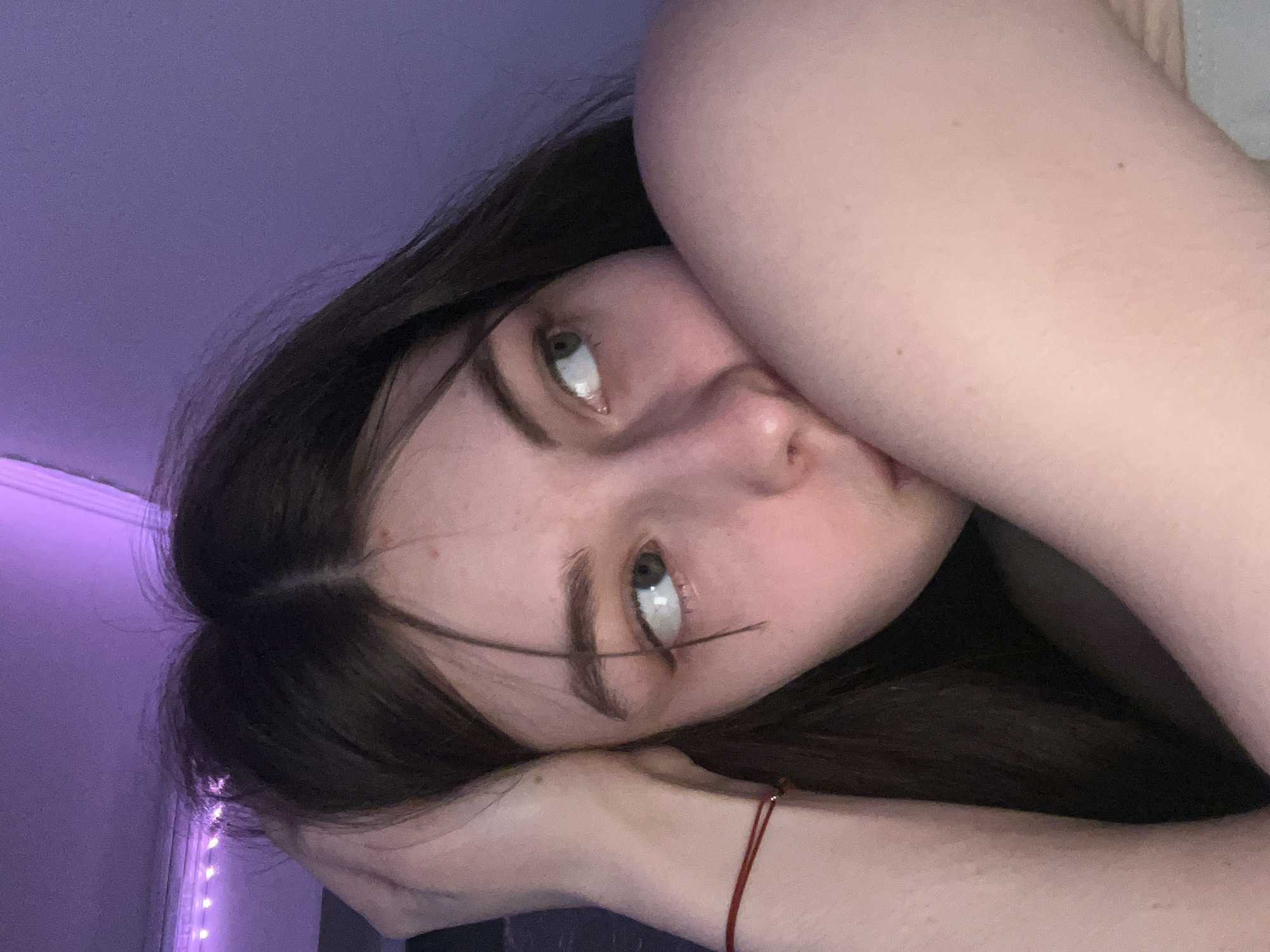 A young woman with dark hair is lying on her side, her face turned towards the viewer. Her arms are bent and positioned near her face. The background appears to be a room with purple lighting. A strand of hair falls across her face, specifically across her eyebrow and eye. A red string bracelet is visible on one of her wrists. The location is unknown and cannot be determined based on the image. There is no indication that the location is Saint Petersburg, Russia.