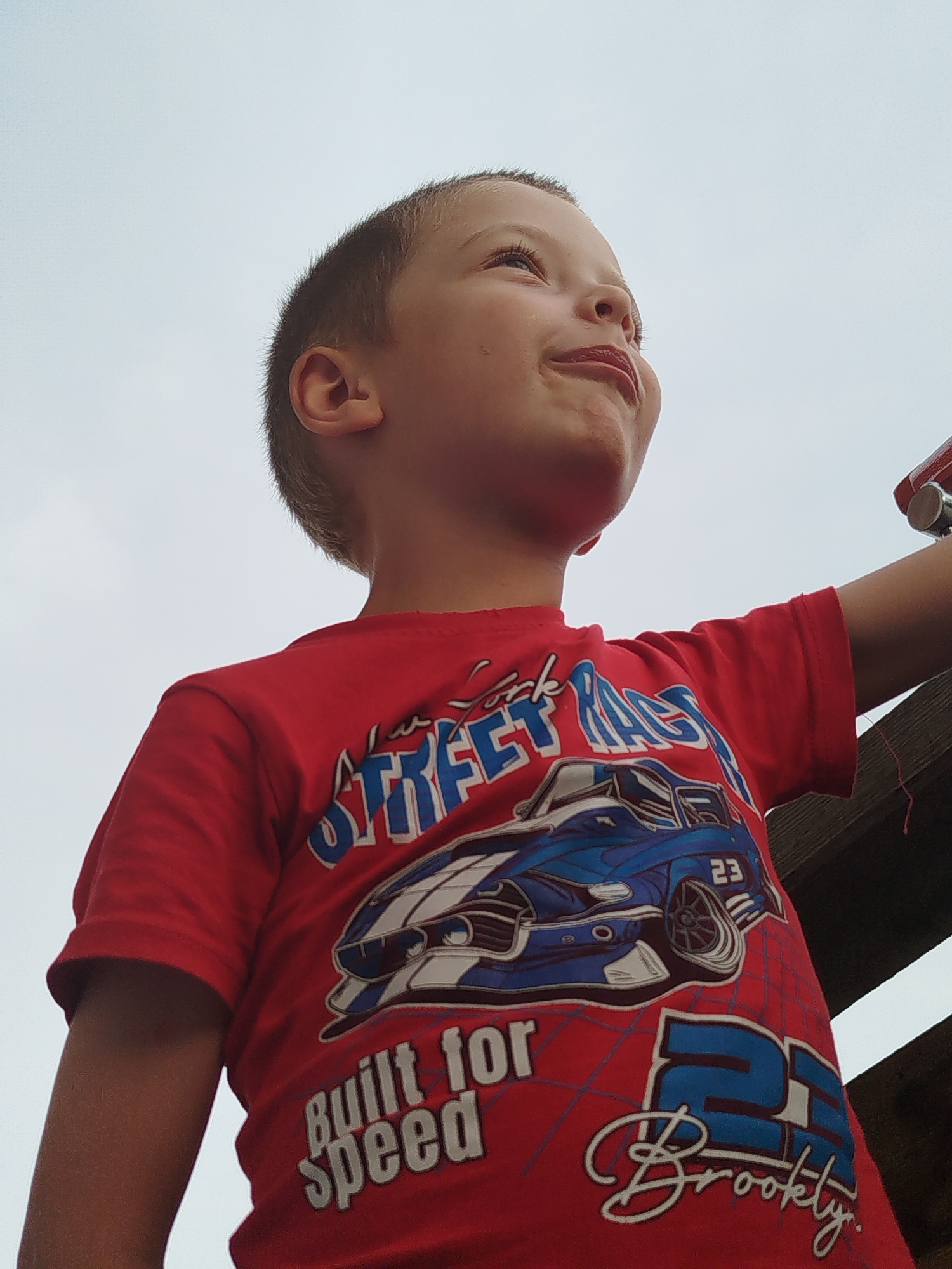 A low-angle, medium shot captures a young boy looking upwards with a slight smile against a pale, overcast sky. He is wearing a red t-shirt with a graphic print. The t-shirt features a blue and white race car with the number 23 on it, along with the text "STREET RACE" and "Built for Speed". Below the car graphic, the number 25 and the word "Brookly" are printed in a stylized font. The boy's right arm is extended upwards and slightly bent, and a wooden structure is visible to the right, partially obscuring the background. No specific location in Zavodoukovsk, Russia, can be identified from the visual information provided.
