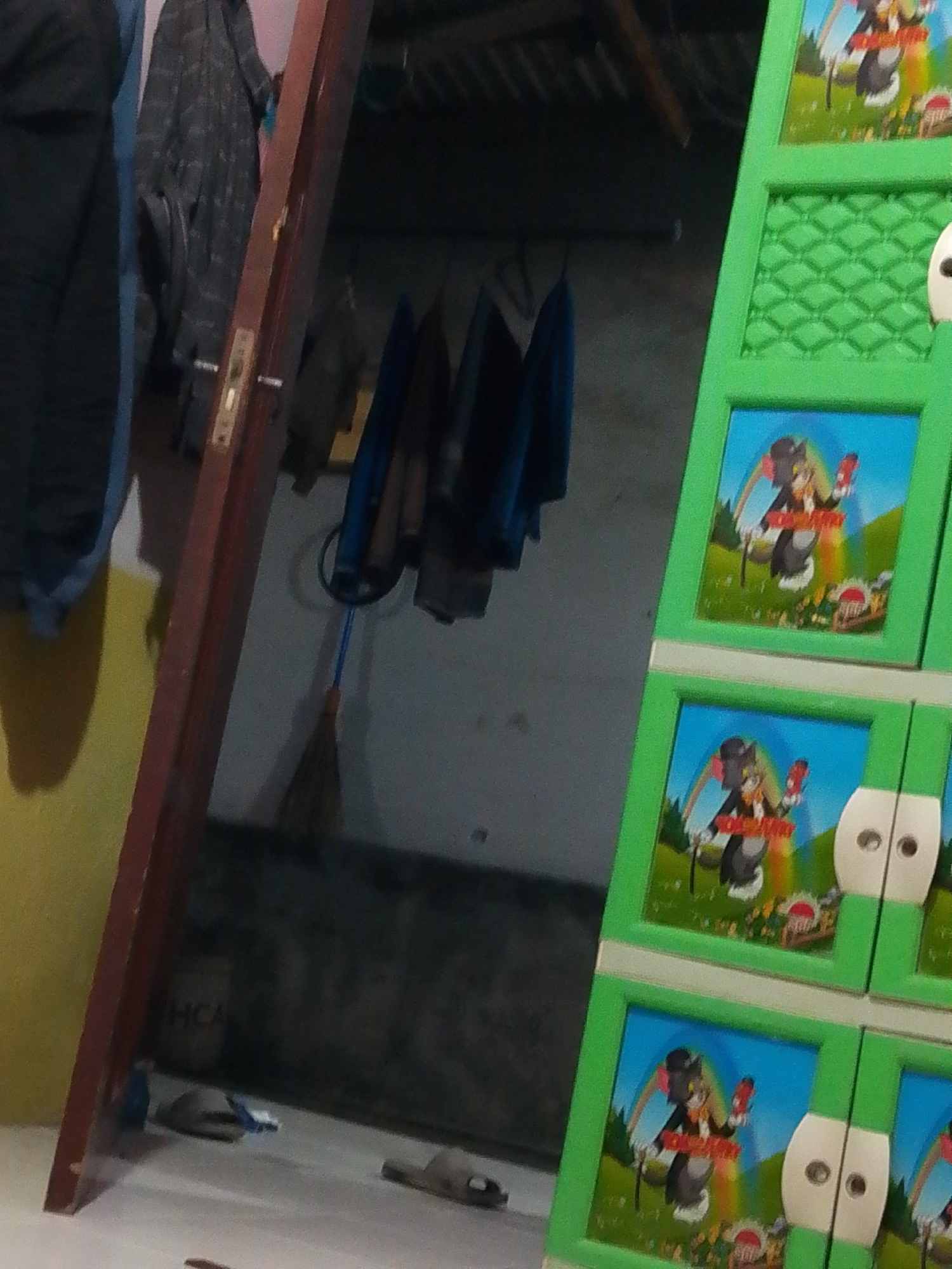This image shows a cluttered interior space. A tall, dark red wooden doorframe dominates the left side of the frame. To the right of the doorframe is a bright green, multi-tiered storage unit with patterned doors. Each door features a cartoon image of a cat character dressed in a hat and suit, holding a red object, with a rainbow and landscape in the background. Several garments, including what appear to be jackets and shirts, are hanging on hooks or a rack behind the doorframe. On the floor, near the bottom of the frame, are a pair of grey slippers and what looks like a small blue toy. In the background, the wall is textured and a portion of a ceiling with wooden beams is visible. The scene appears to be located within a residential dwelling in Bohar, Indonesia, likely a storage area or a room that doubles as such.